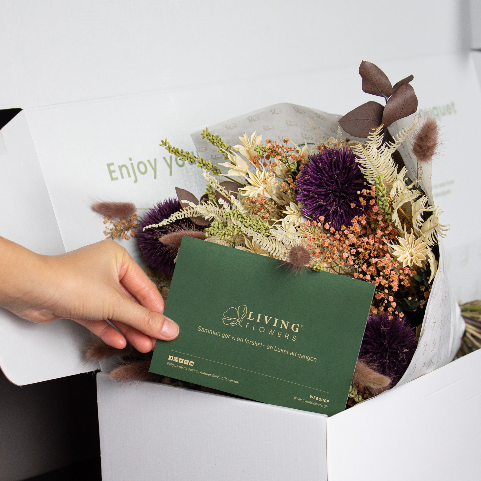 I en eksklusiv hvid Êske ligger en buket, mens en hÄnd lÊgger et mÞrkegrÞnt kort ved.