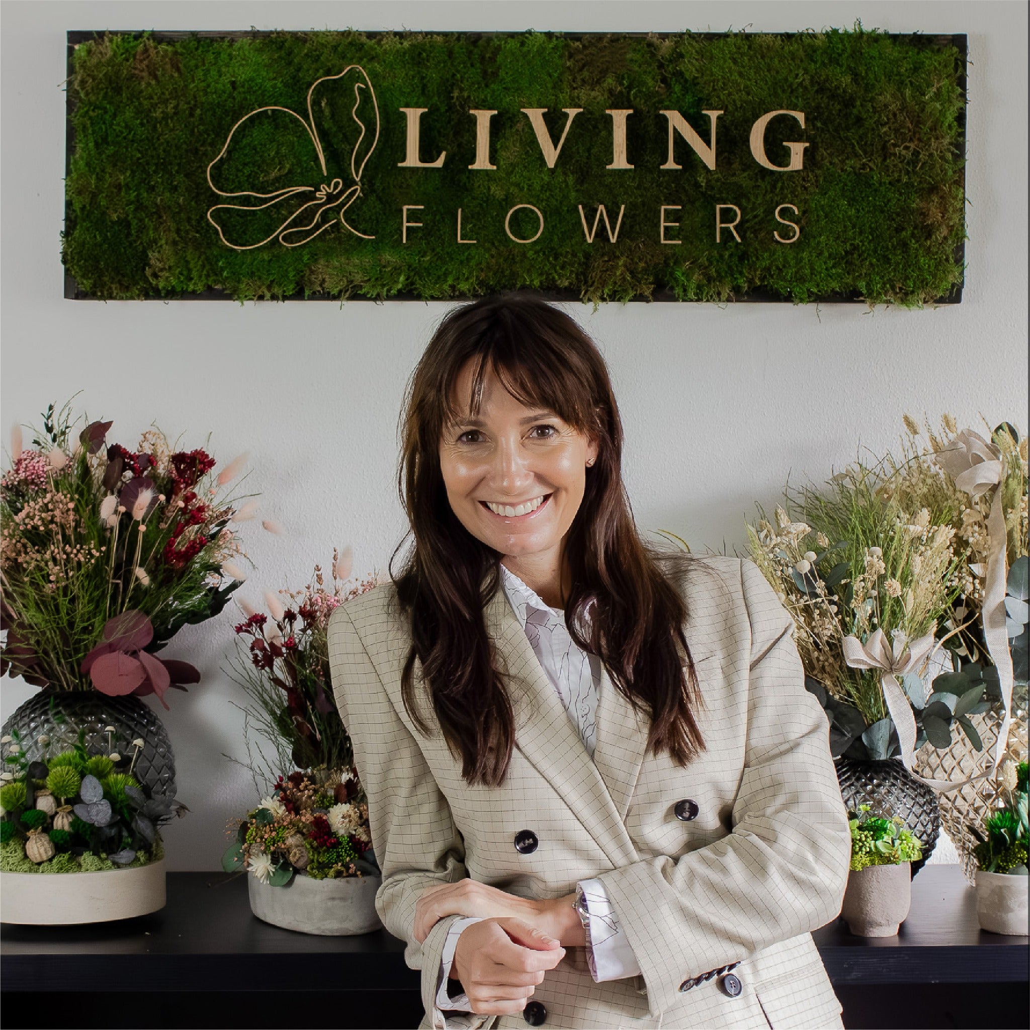 Grundlæggeren af Living Flowers står smilende foran en reol med langtidsholdbare dekorationer og blomsterbuketter. Over hende hænger et skilt med teksten „Living Flowers“.