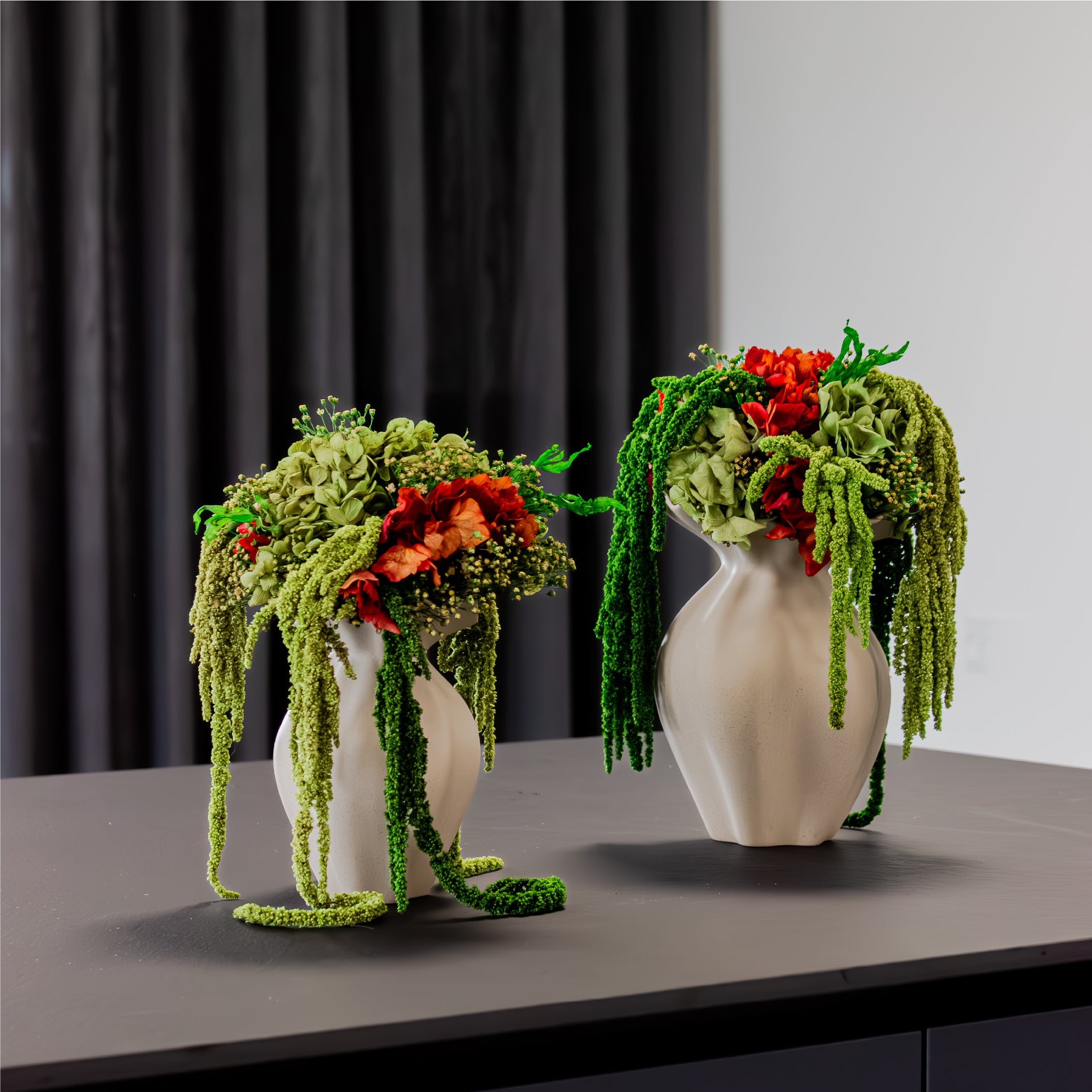 På et bord står to detaljeret bundne grøn-orange blomsterbuketter med hængende elementer i sandfarvede keramikkvaser.