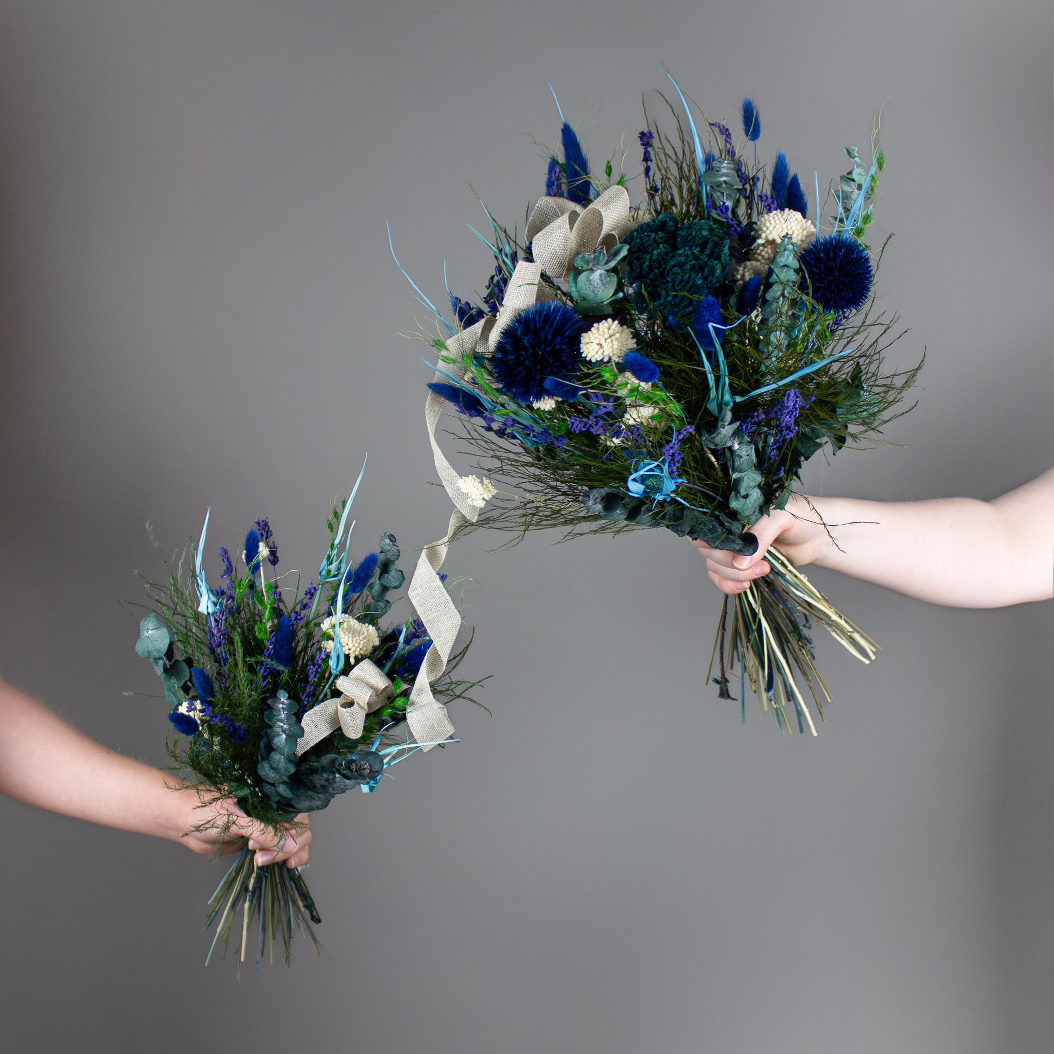 To hænder holder hver sin buket i blå og hvide nuancer: En mindre børnebuket er forbundet med et bånd til en større buket i samme design.