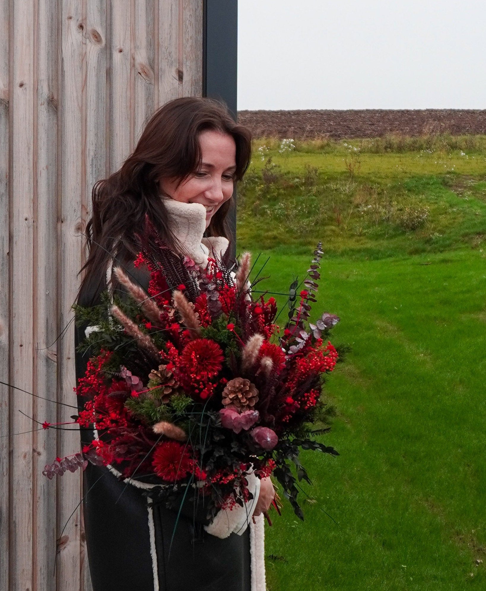 En smilende kvinde i vinterfrakke holder en fyldig buket i julefarver i armene.