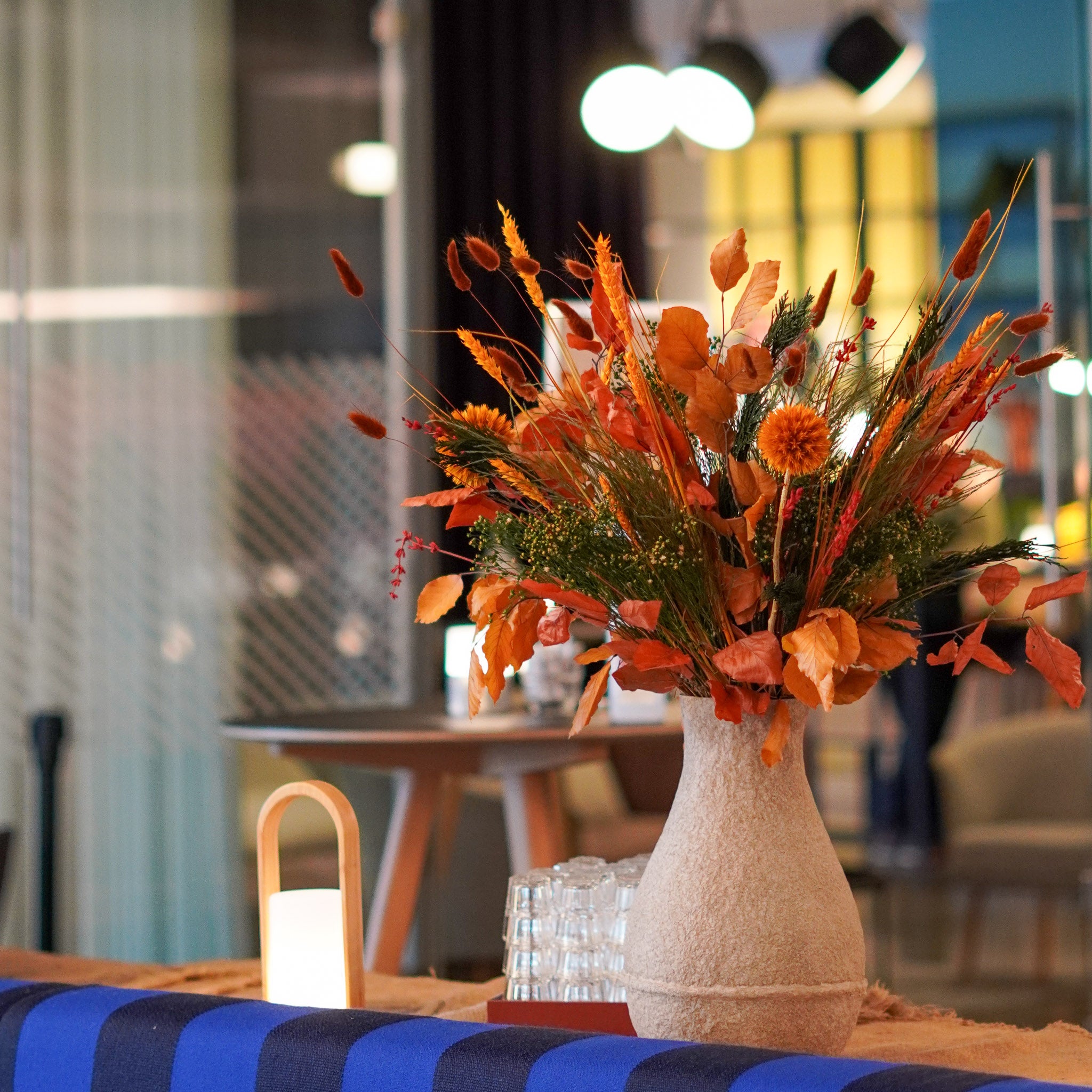 En efterårsbuket i kraftige orange og grønne nuancer står i en beige vase på et dækket bord i et moderne indrettet rum.