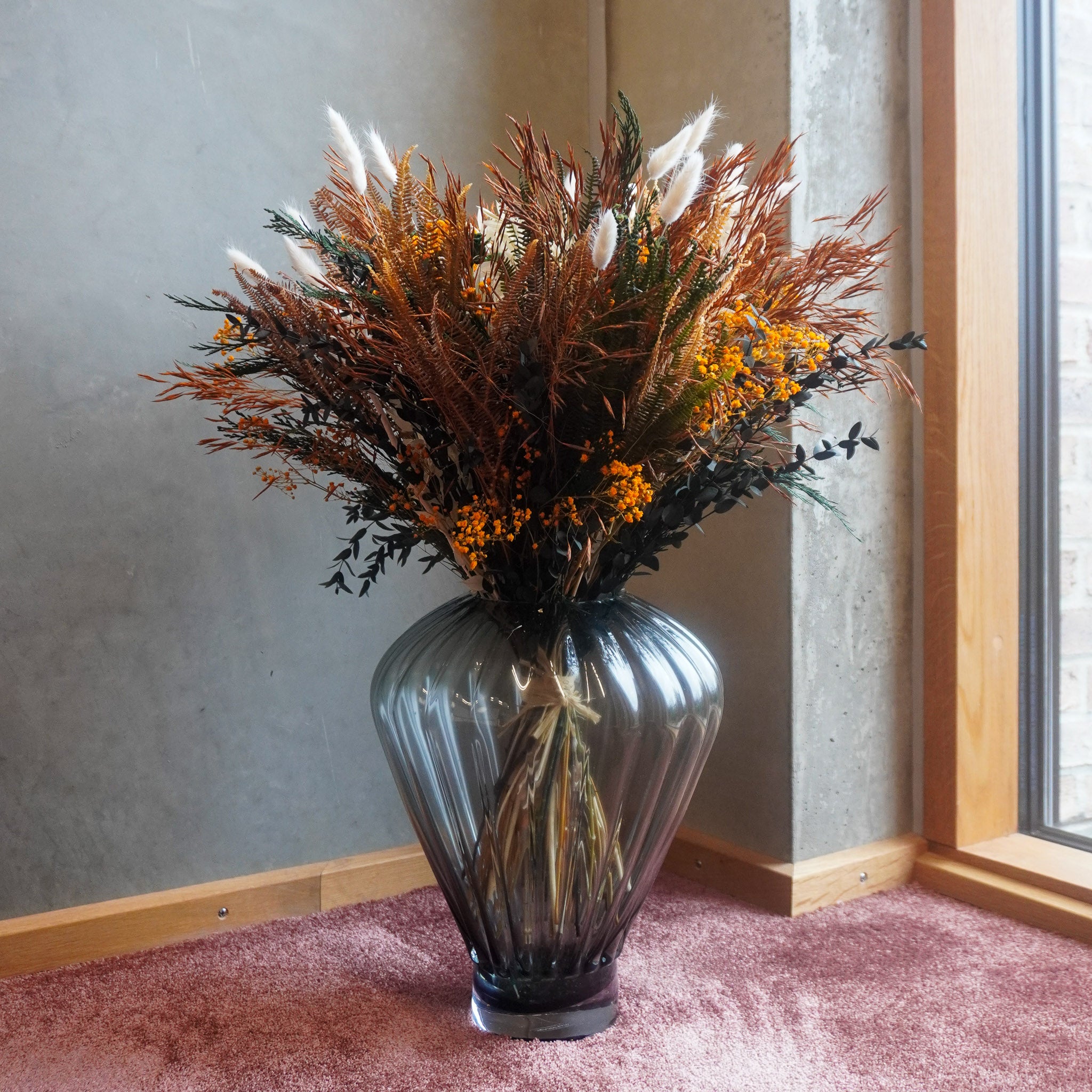 I et hjÞrne af et vÊrelse stÄr en fyldig blomsterbuket i varme efterÄrsfarver i en geometrisk glasvase pÄ gulvet.