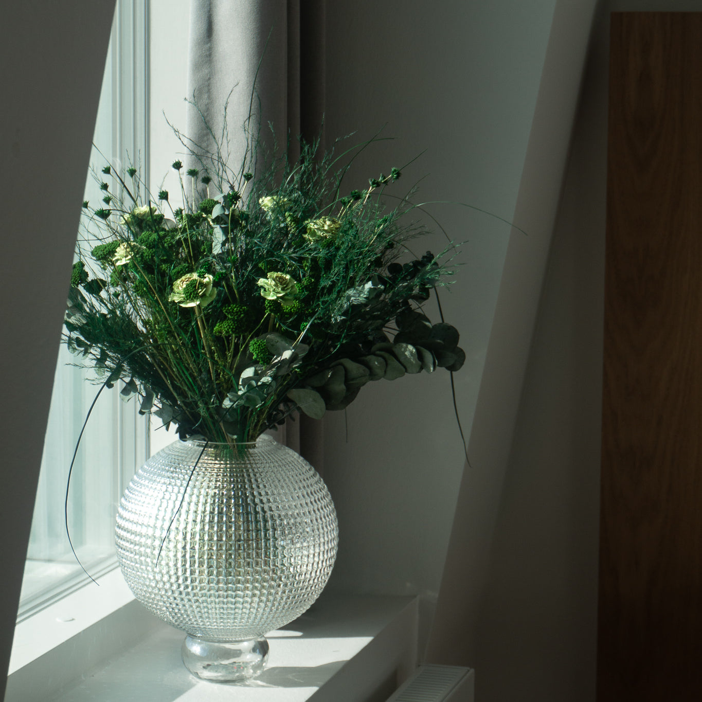 Bukket med langtidsholdbare blomster i stærke grønne nuancer står i en glasvase på en vindueskarm.