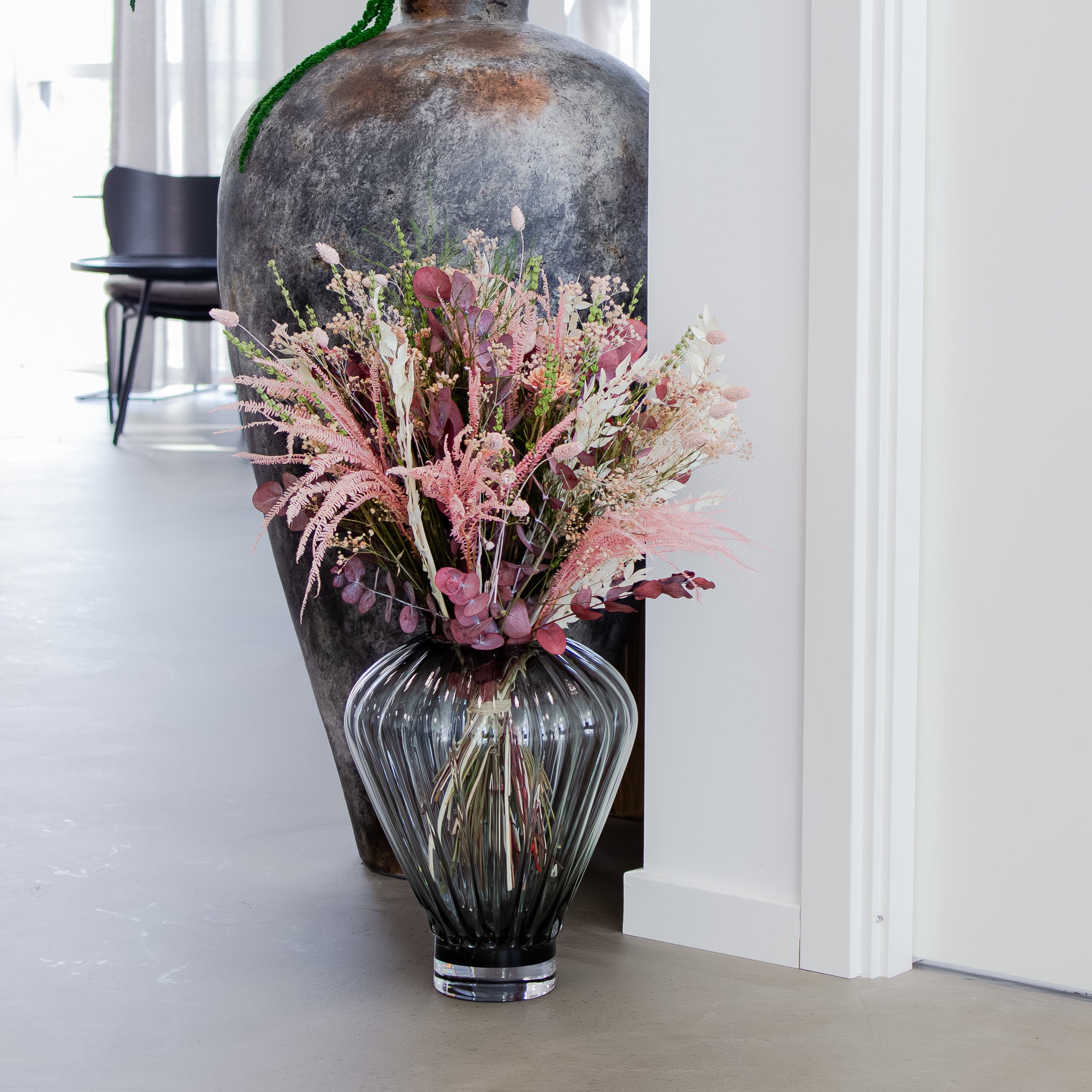 I et hjørne af et værelse står en stor, rillet glasvase med en blomsterbuket i sarte rosa nuancer.