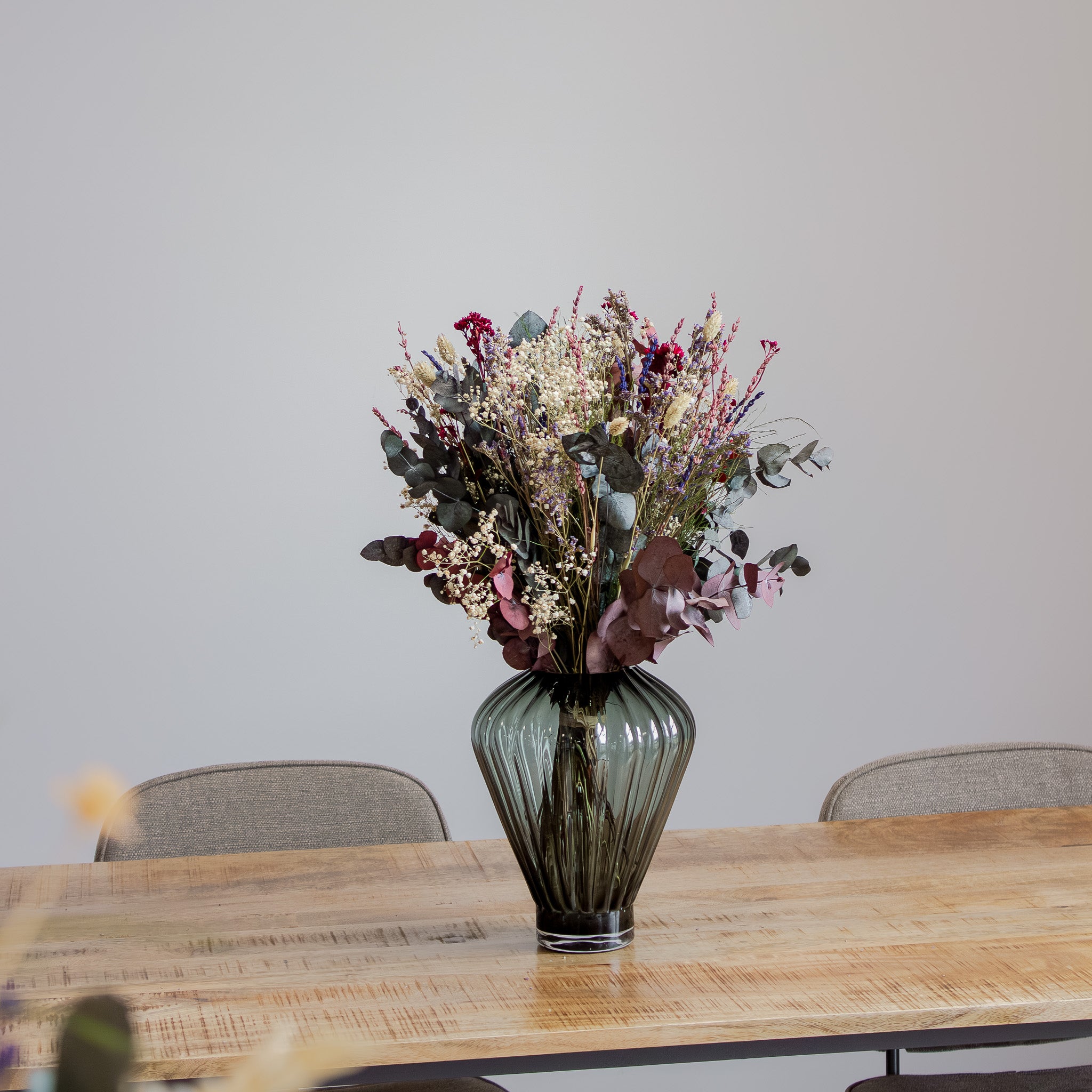 En vild, naturlig buket med violette, blå og røde accenter står i en rillet glasvase på et træbord.