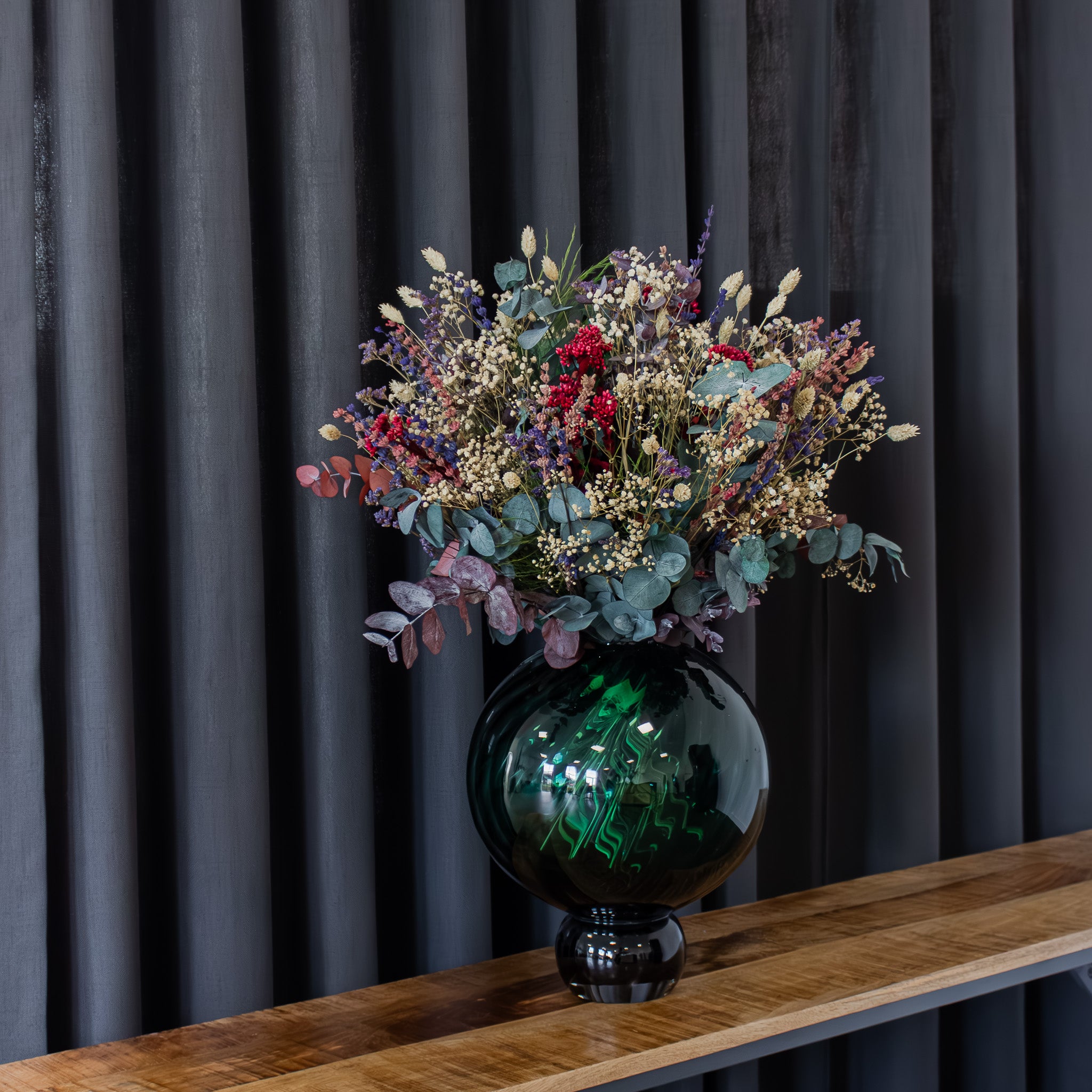 En fyldig, naturlig buket med lilla, blå og røde nuancer står i en rund glasvase.