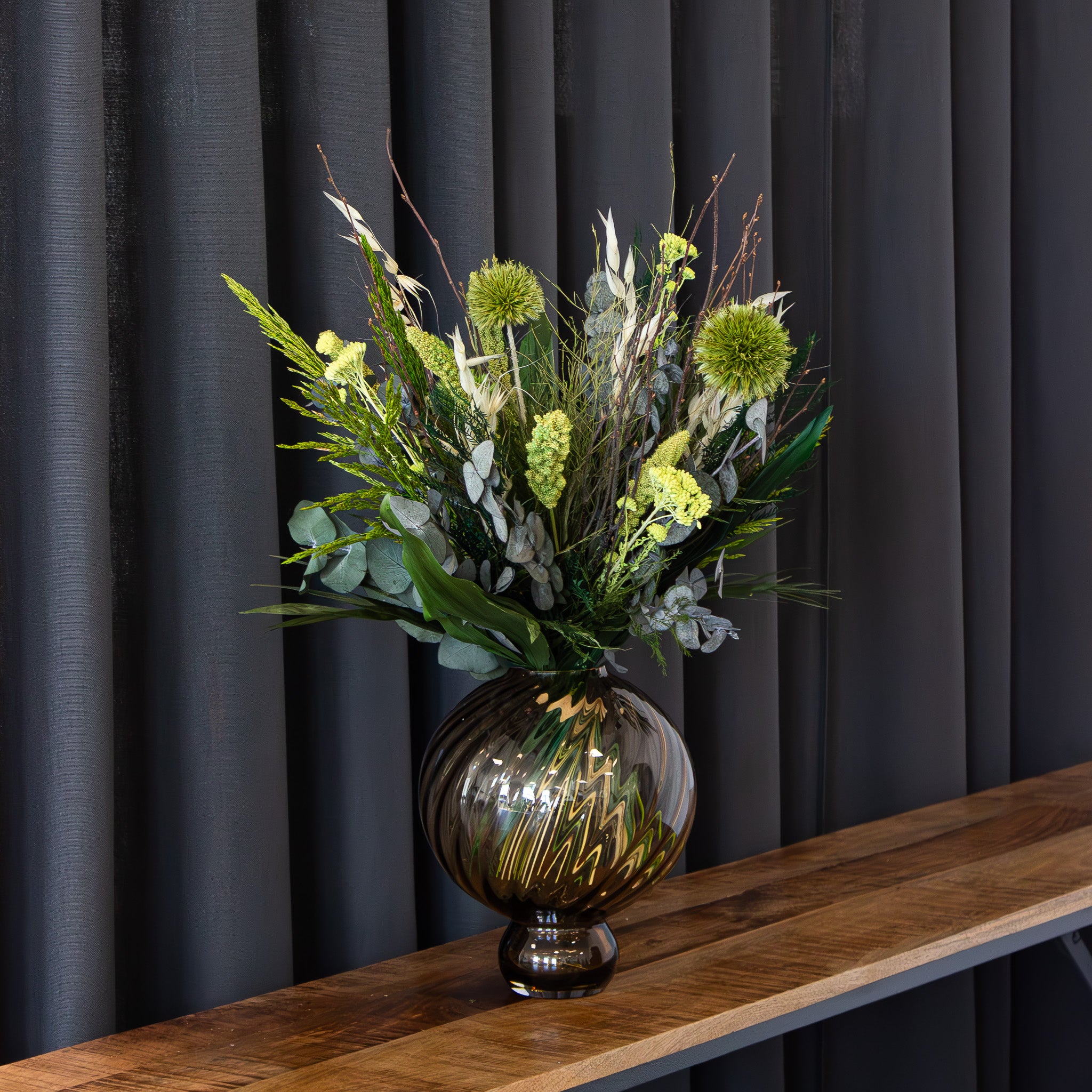 I en rund glasvase står en blomsterbuket i dybe grønne nuancer.