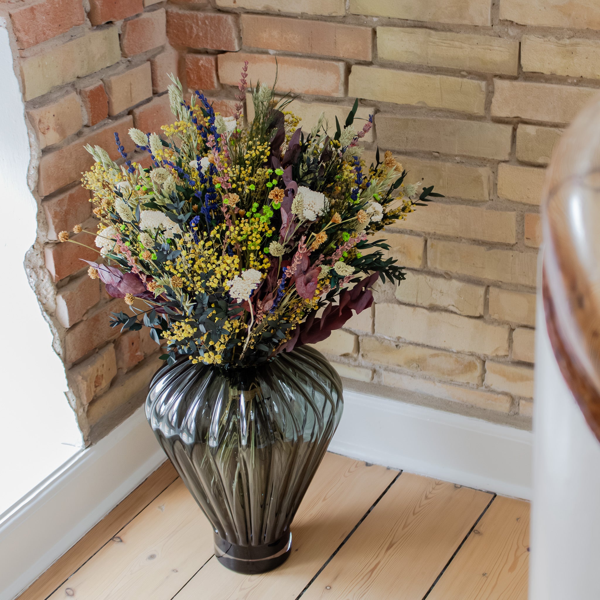 I et hjørne af et værelse står en stor glasvase med en farverig blomsterbuket