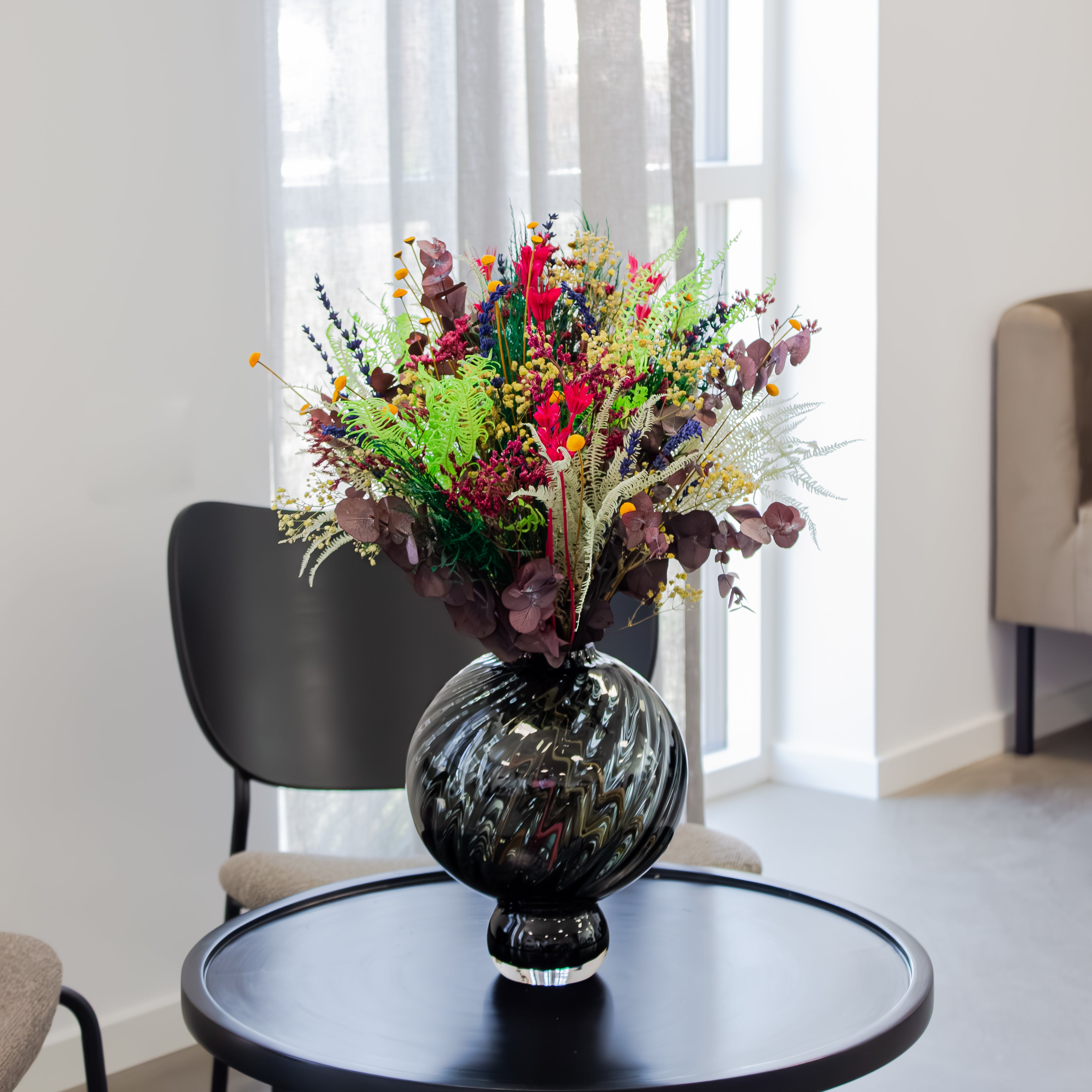 En stor, farverig blomsterbuket med grønne, røde og violette nuancer står i en rillet, buet glasvase på et rundt, sort bord i et moderne rum.