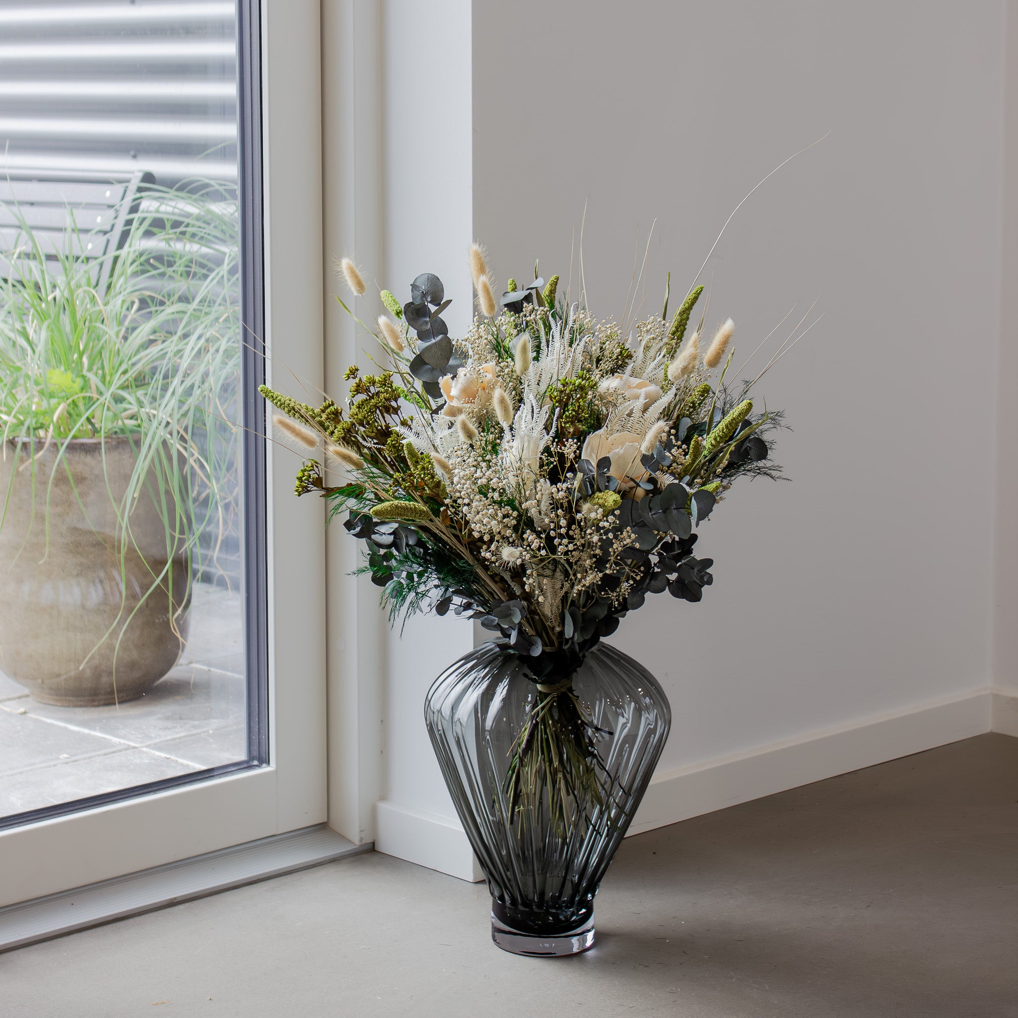 Naturlig bukket i hvide og grønne nuancer står i en stor, geometrisk glasvase placeret på gulvet i et hjørne af rummet ved et vindue.
