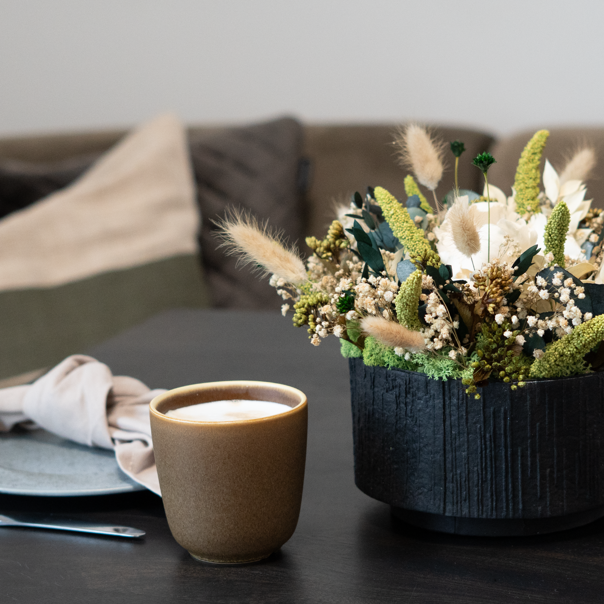 Dekoration af langtidsholdbare blomster i en sort papbakke på et bord ved siden af en kop.