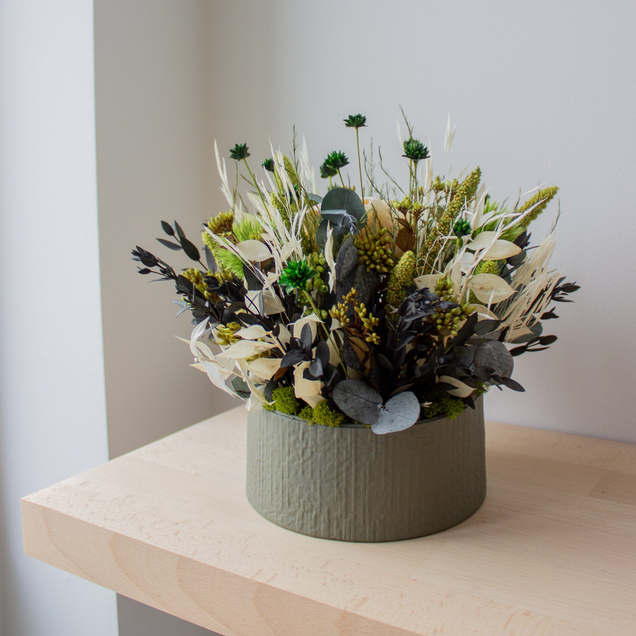 En blomsterdekoration med langtidsholdbare blomster i grønne og cremefarvede nuancer står i en skål på en lys træhylde.