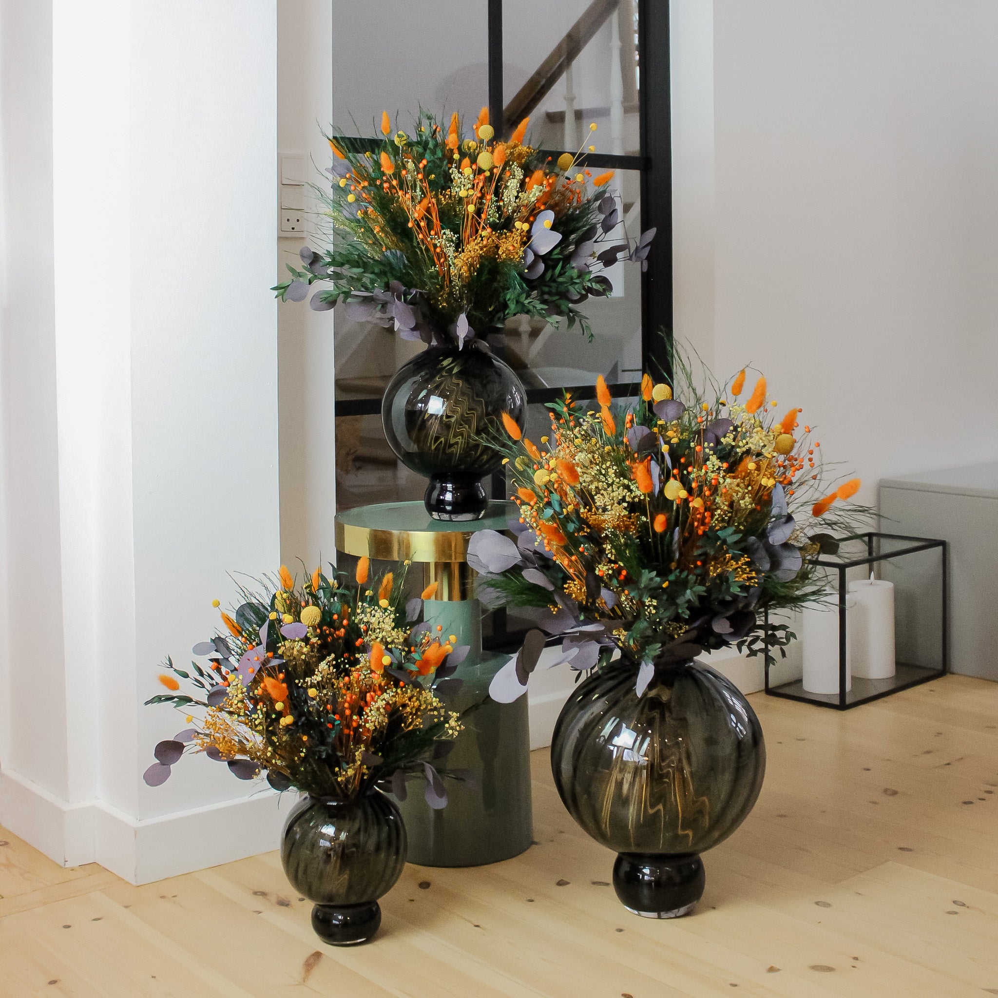 Tre buketter i forskellige størrelser står i farvekoordinerede, kugleformede vaser – hver vase tilpasset størrelsen på den enkelte buket – arrangeret på gulvet og en piedestal i et lyst og moderne rum.