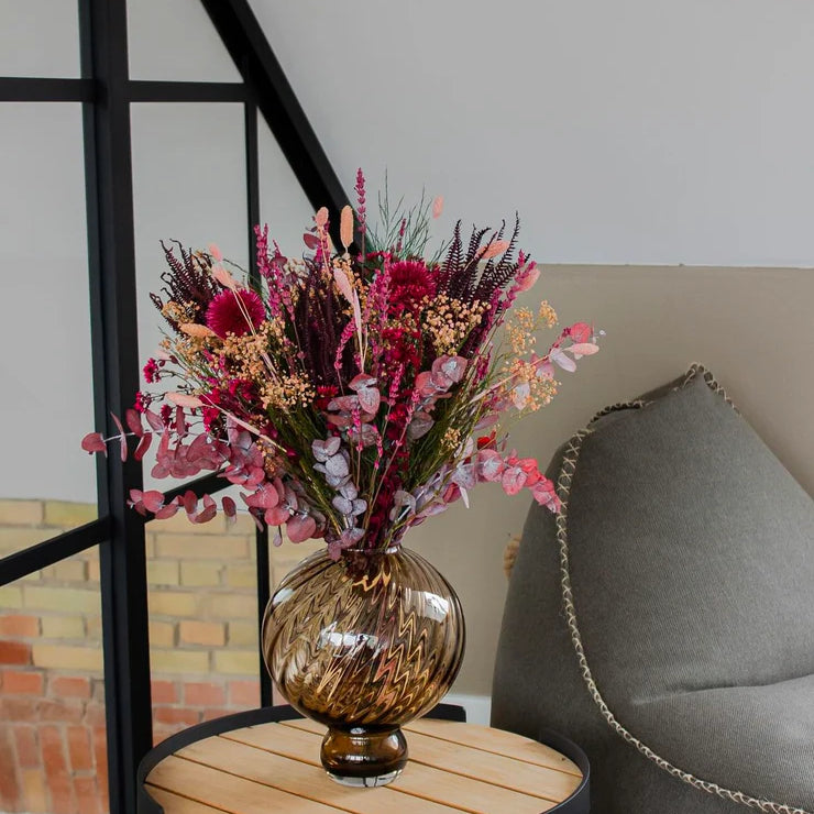 En elegant blomsterbuket i rosa-røde nuancer står i en rippet vase på et sidebord.