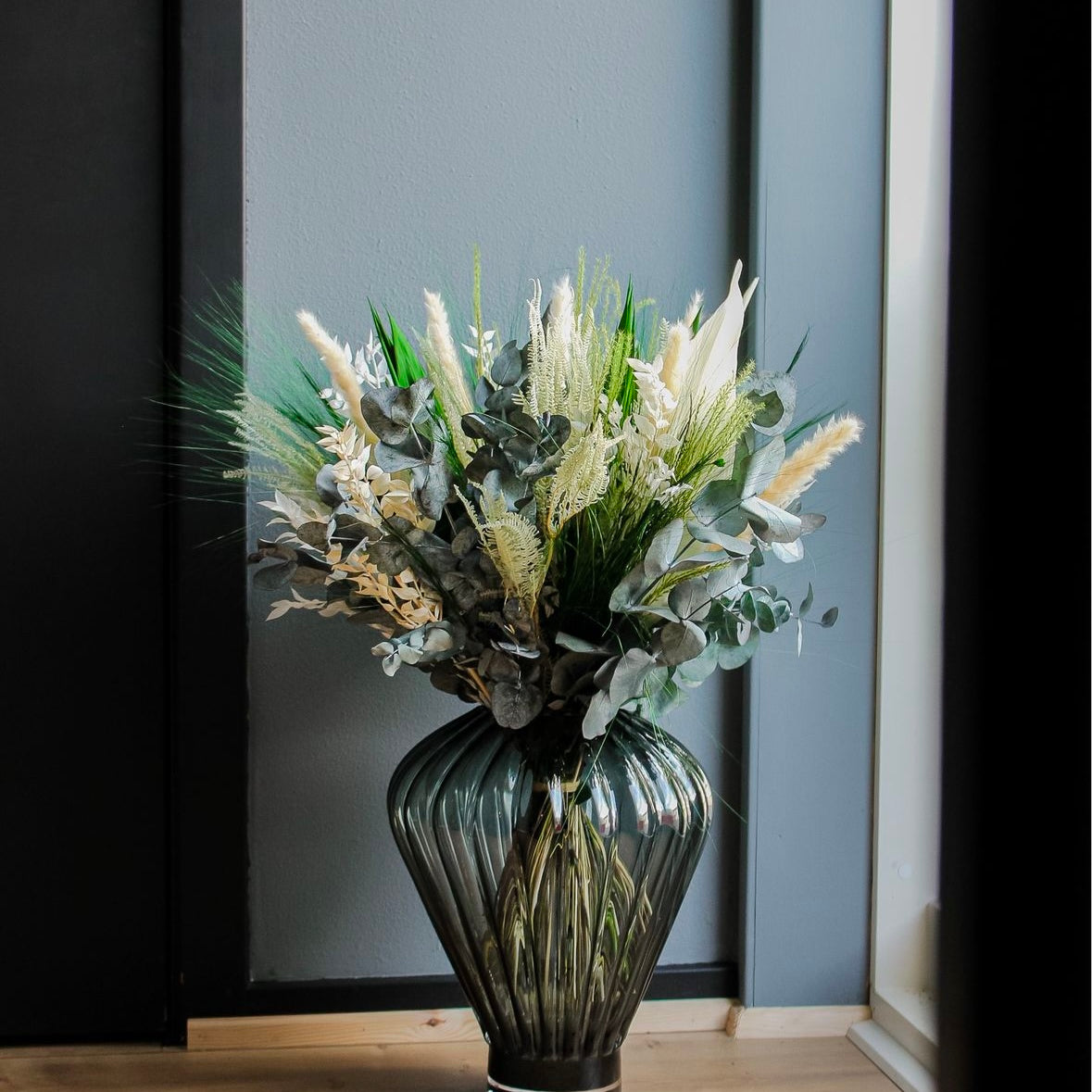 En grøn-hvid buket står i en stor riflet glasvase på gulvet.
