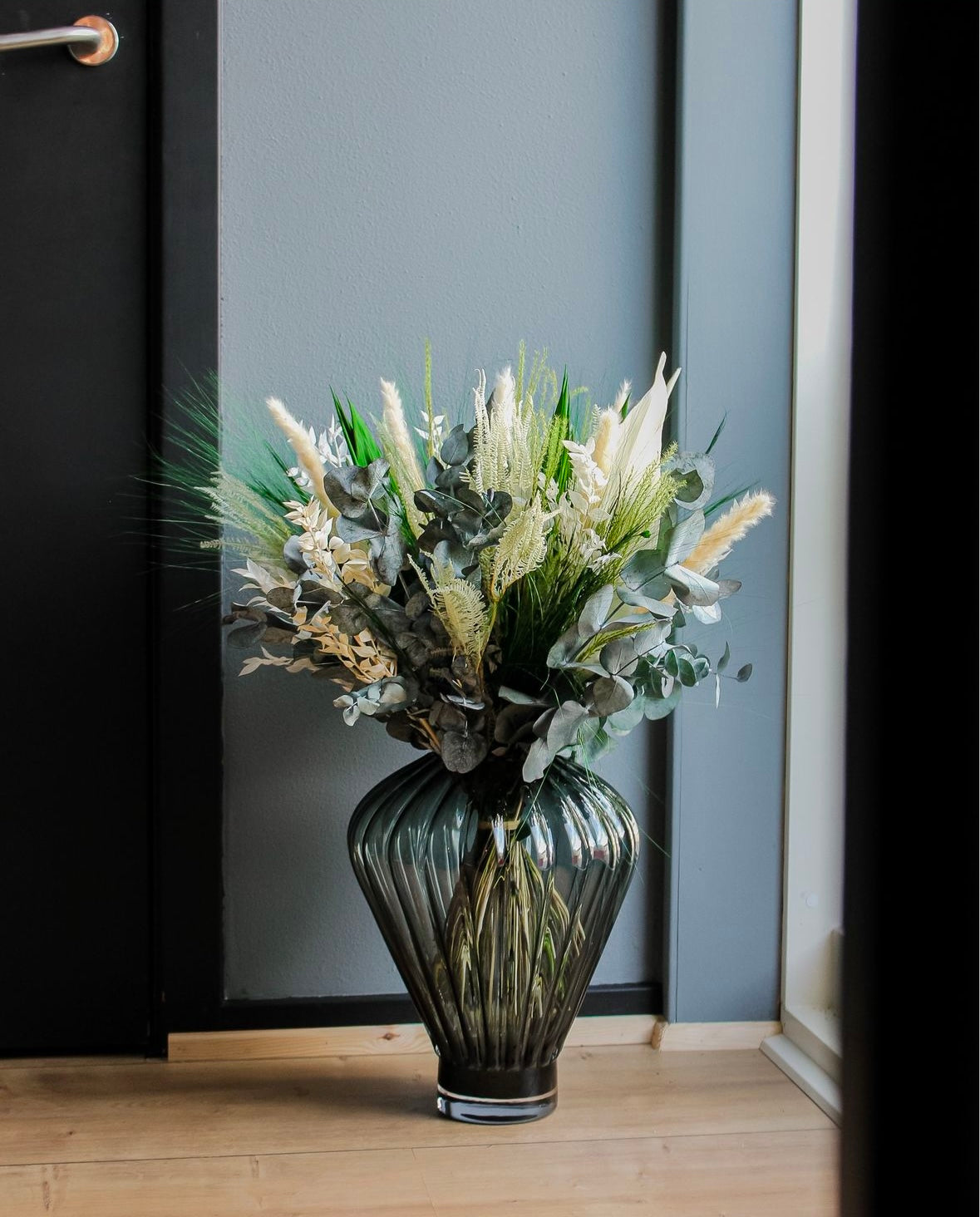 En grøn-hvid buket står i en stor riflet glasvase på gulvet.