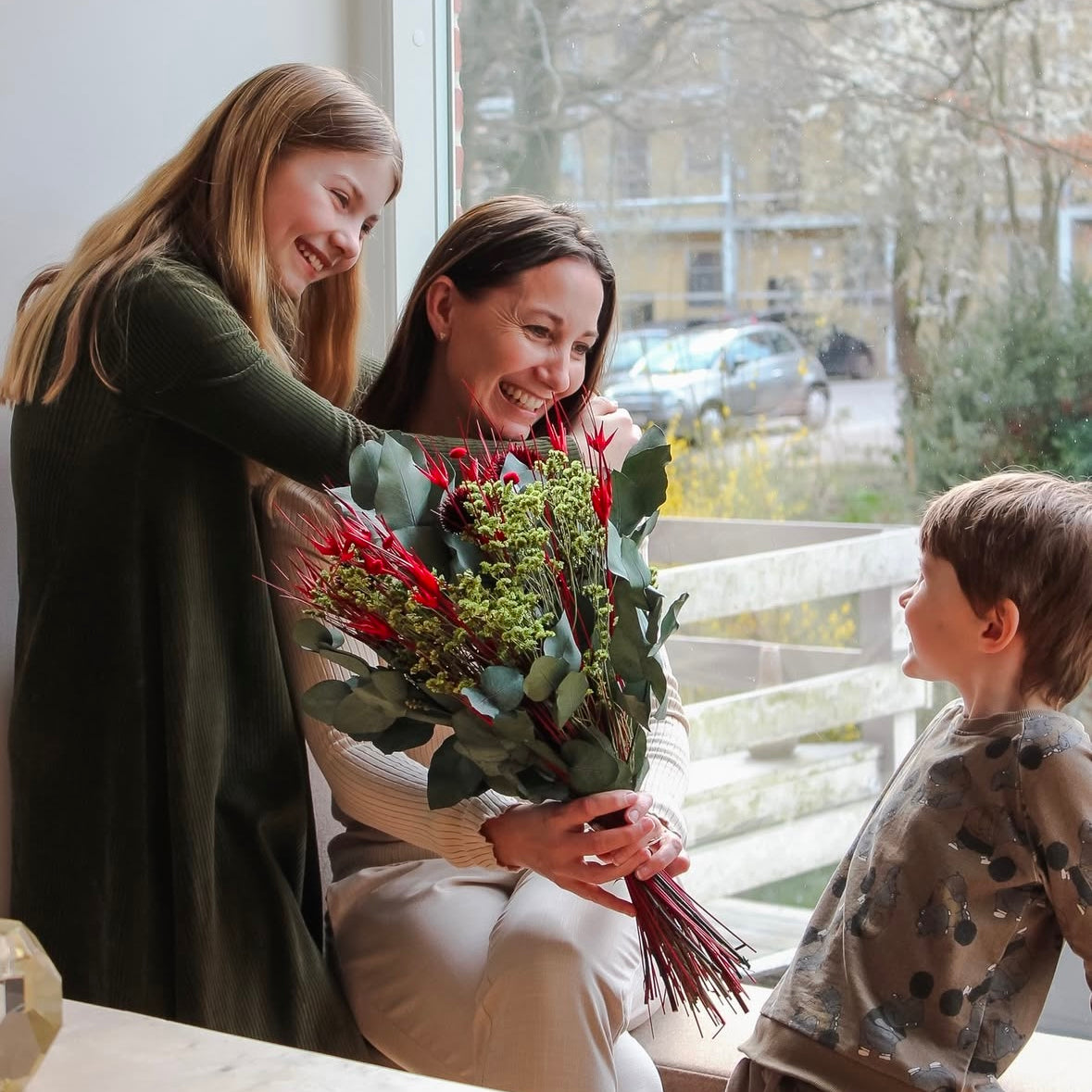 En mor sidder med sine to børn i en stue, alle smiler. Moren holder en grøn-rød buket.