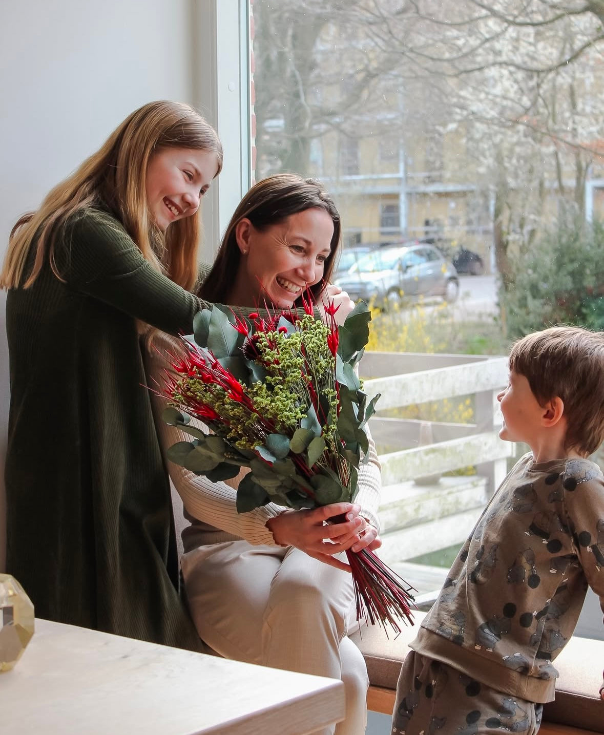En mor sidder med sine to børn i en stue, alle smiler. Moren holder en grøn-rød buket.