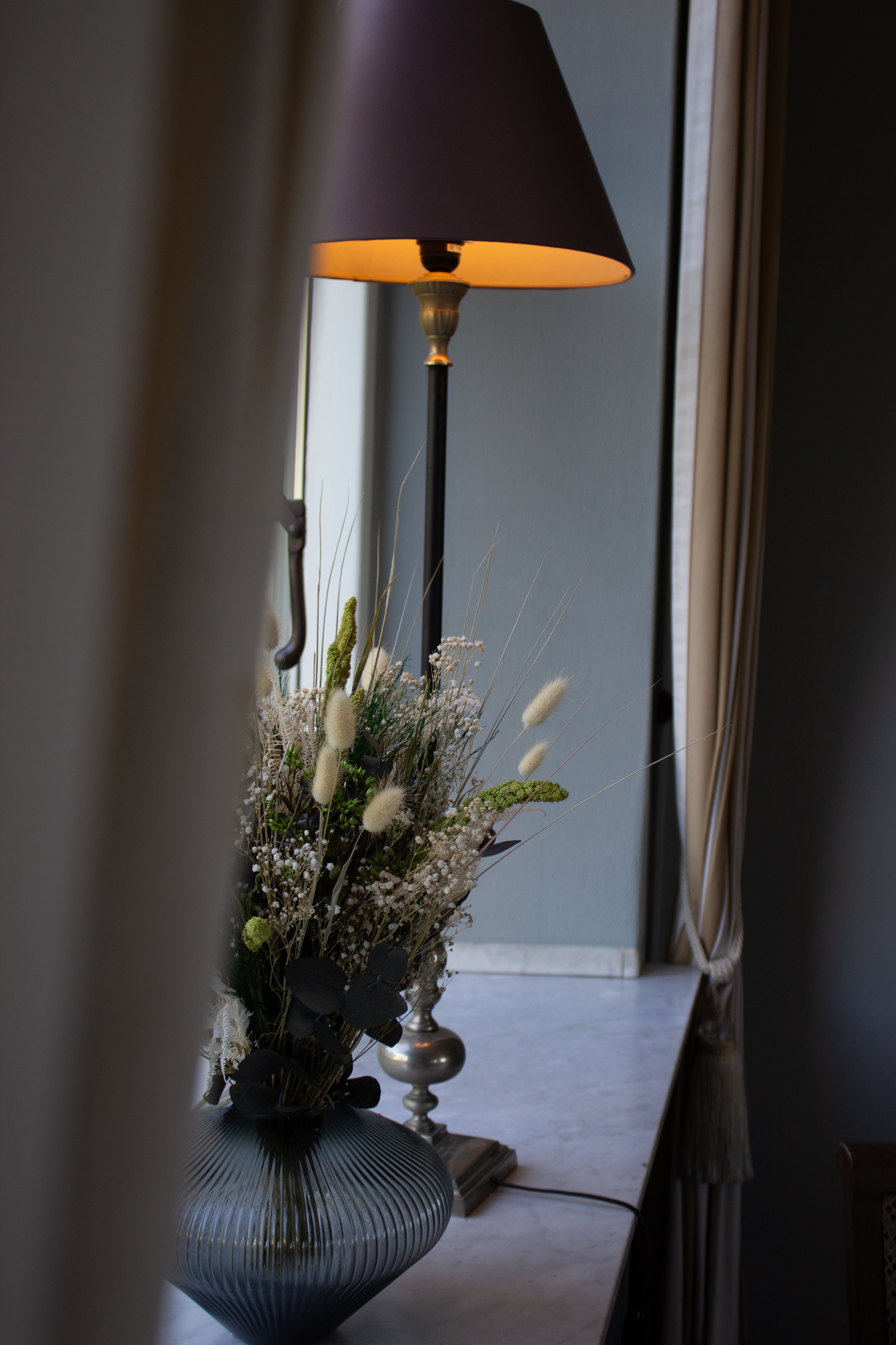 En grøn-hvid buket med langtidsholdbare blomster står i en glasvase i vindueskarmen ved siden af en gammeldags lampe.