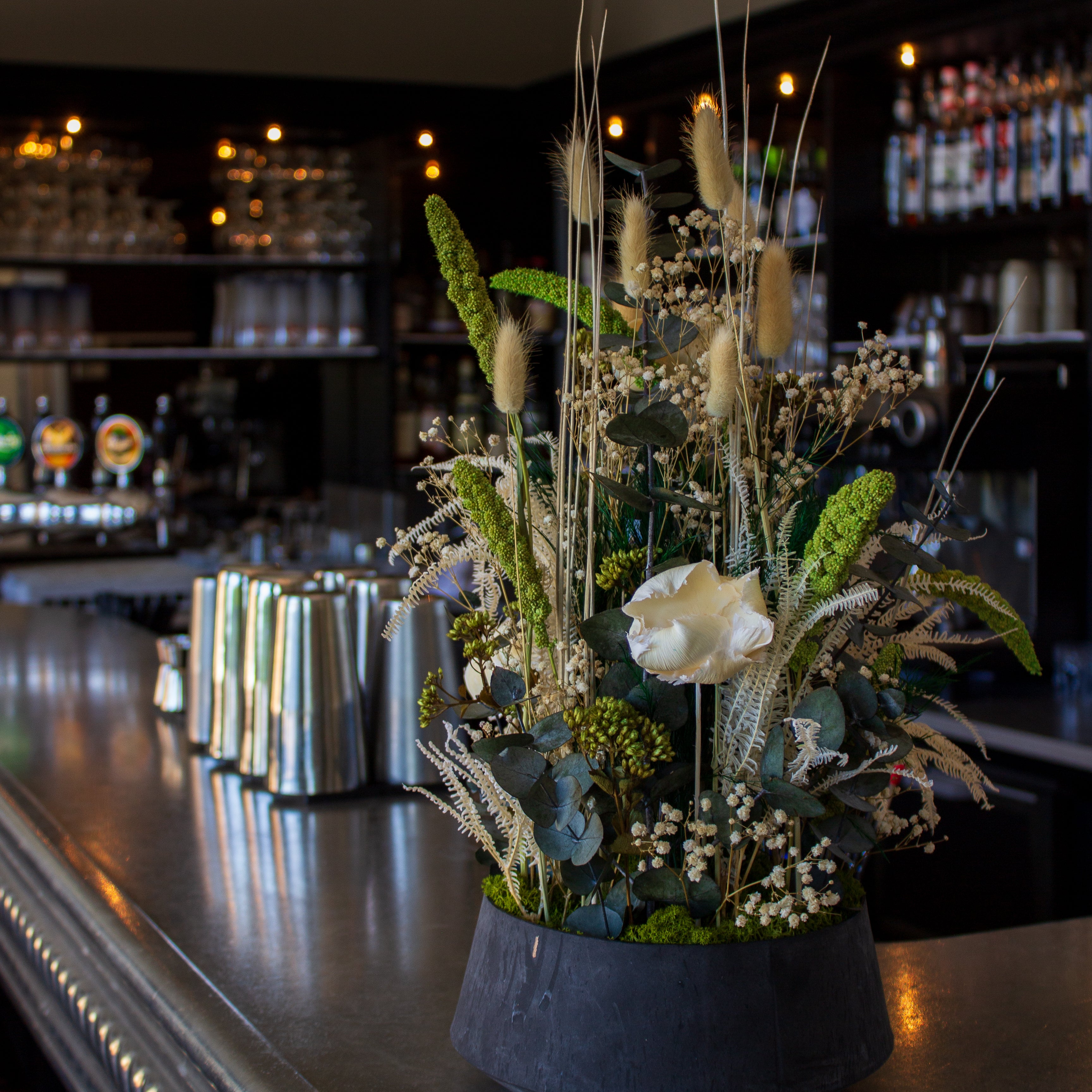 På en bardisk står en imponerende blomsterdekoration i naturlige hvid-grønne nuancer i en skål.