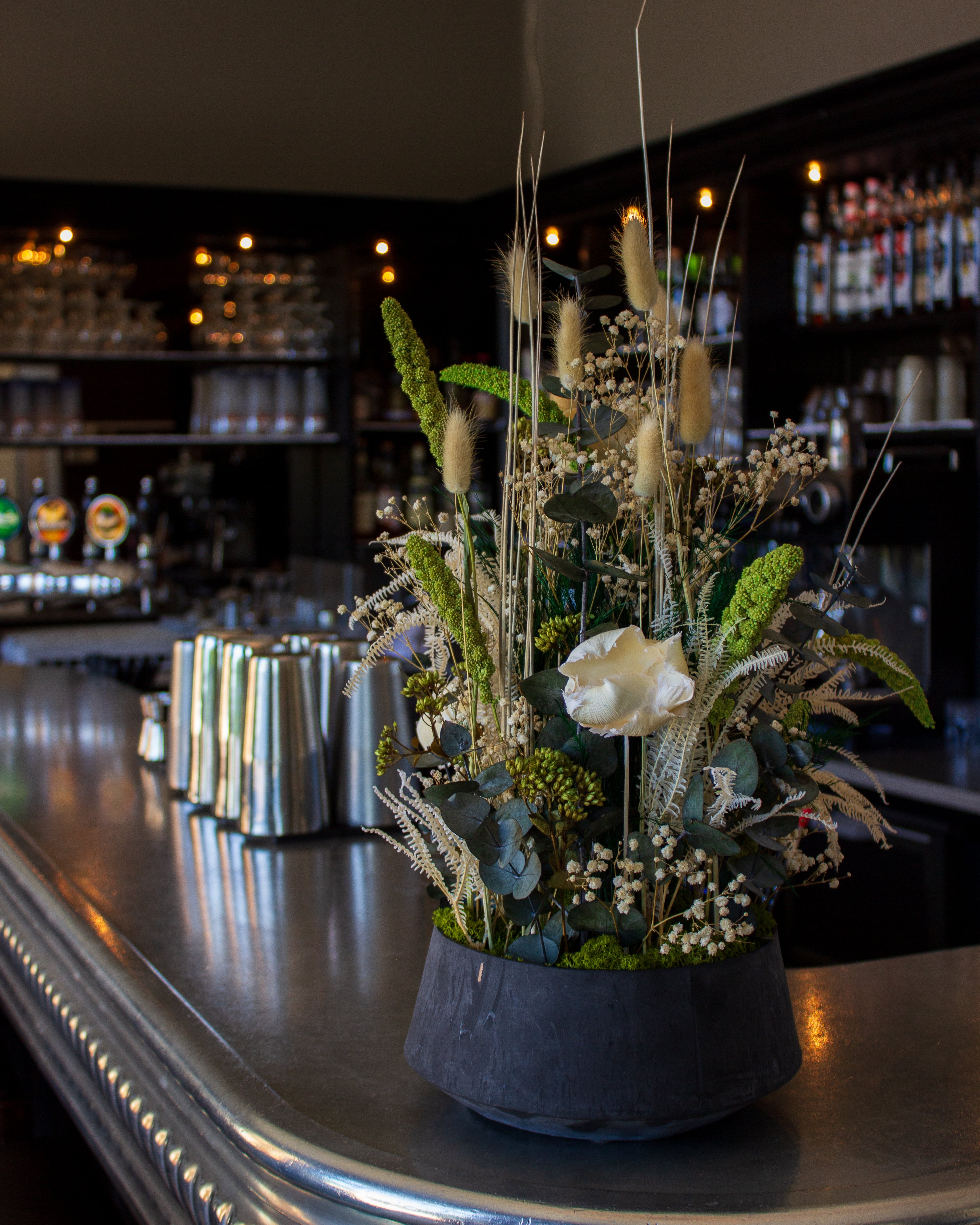 På en bardisk står en imponerende blomsterdekoration i naturlige hvid-grønne nuancer i en skål.