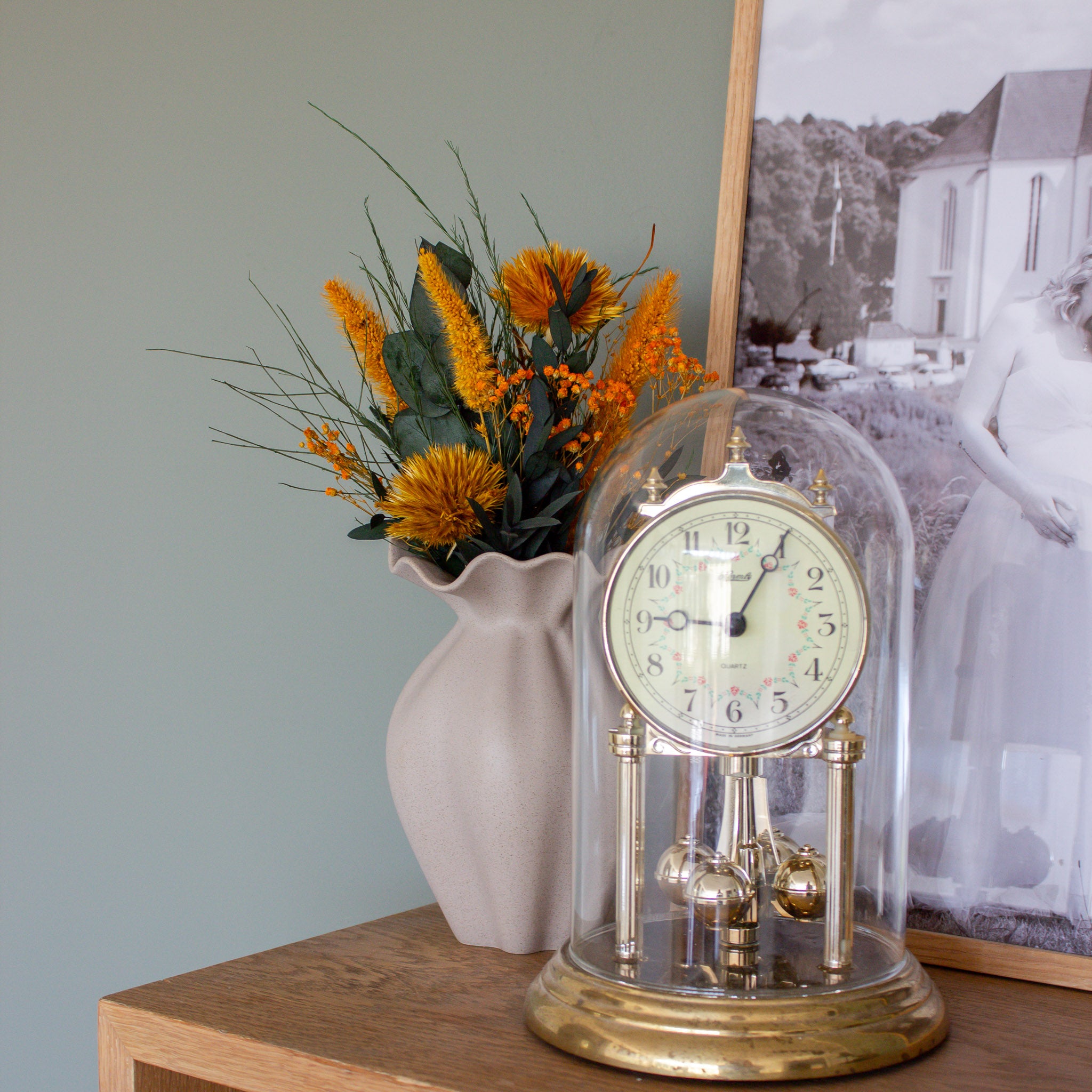 En mini-buket i kraftig orange står i en sandfarvet vase ved siden af et antikt ur på en hylde.