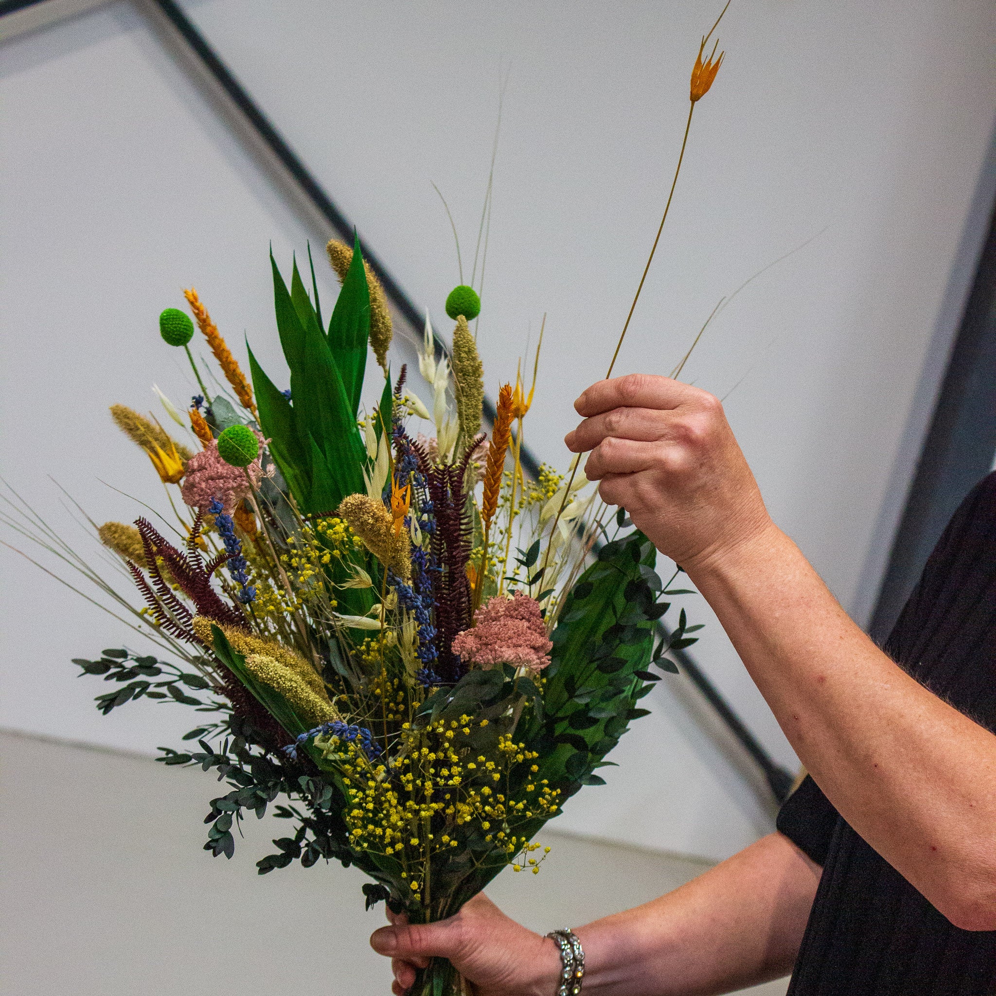 En florist holder en farverig buket i højre hånd og placerer en langtidsholdbar, orange blomst i buketten med venstre hånd.