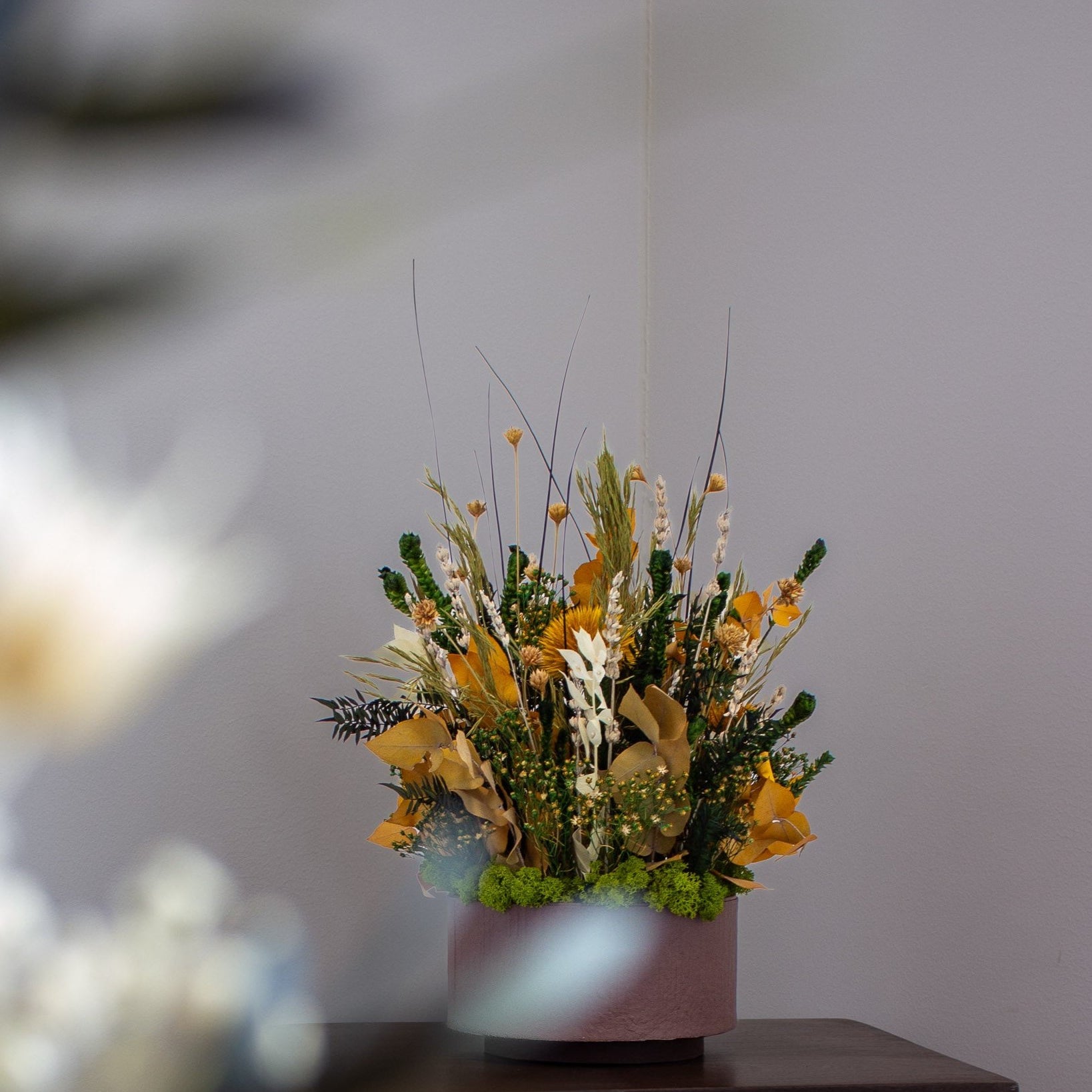 En langtidsholdbar blomsterbuket i efterårsfarver med gule, grønne og naturfarvede toner står i en lyserød beholder på et mørkt træbord foran en grå væg.