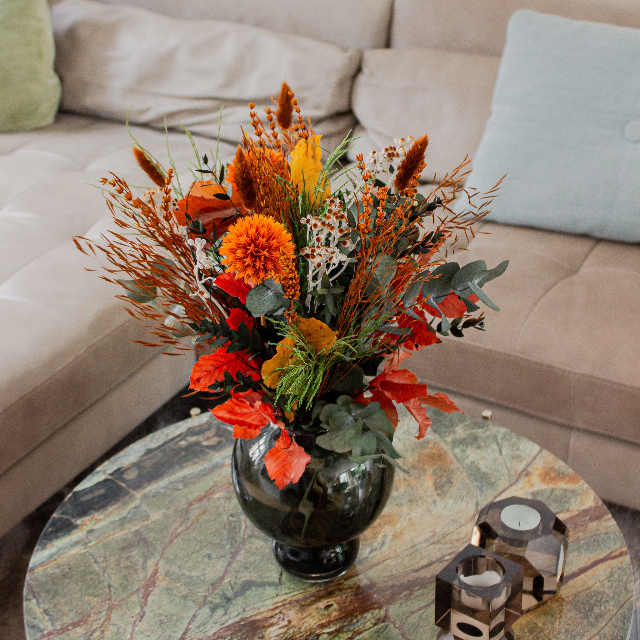 Bukket med langtidsholdbare blomster i varme efterårsfarver står i en mørkegrøn, buet vase på et marmorbord foran en beige sofa.