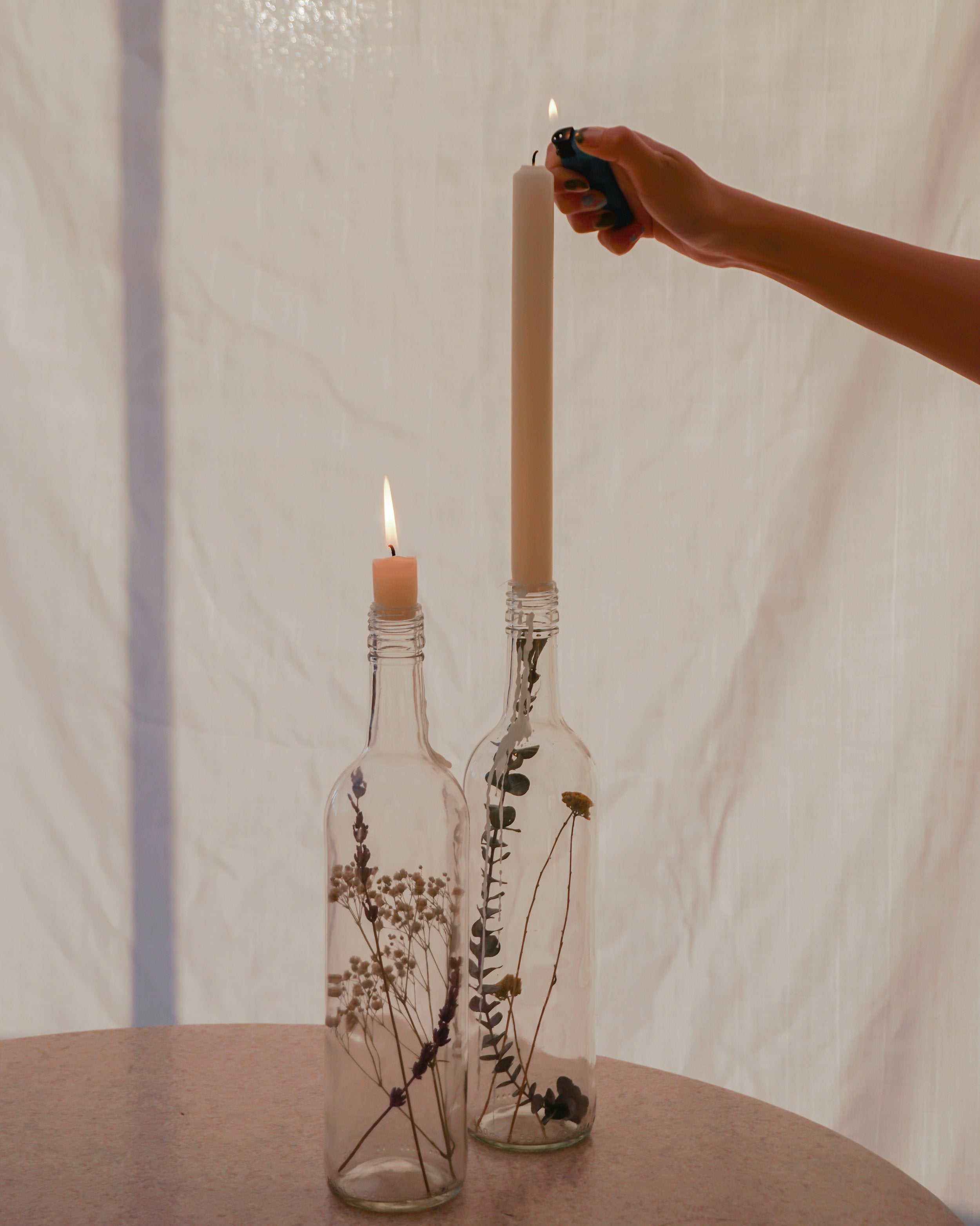 To glasflasker, der er omdannet til lysestager, står på et bord. Flaskerne indeholder tørrede blomster, og en kvinde tænder lysene.