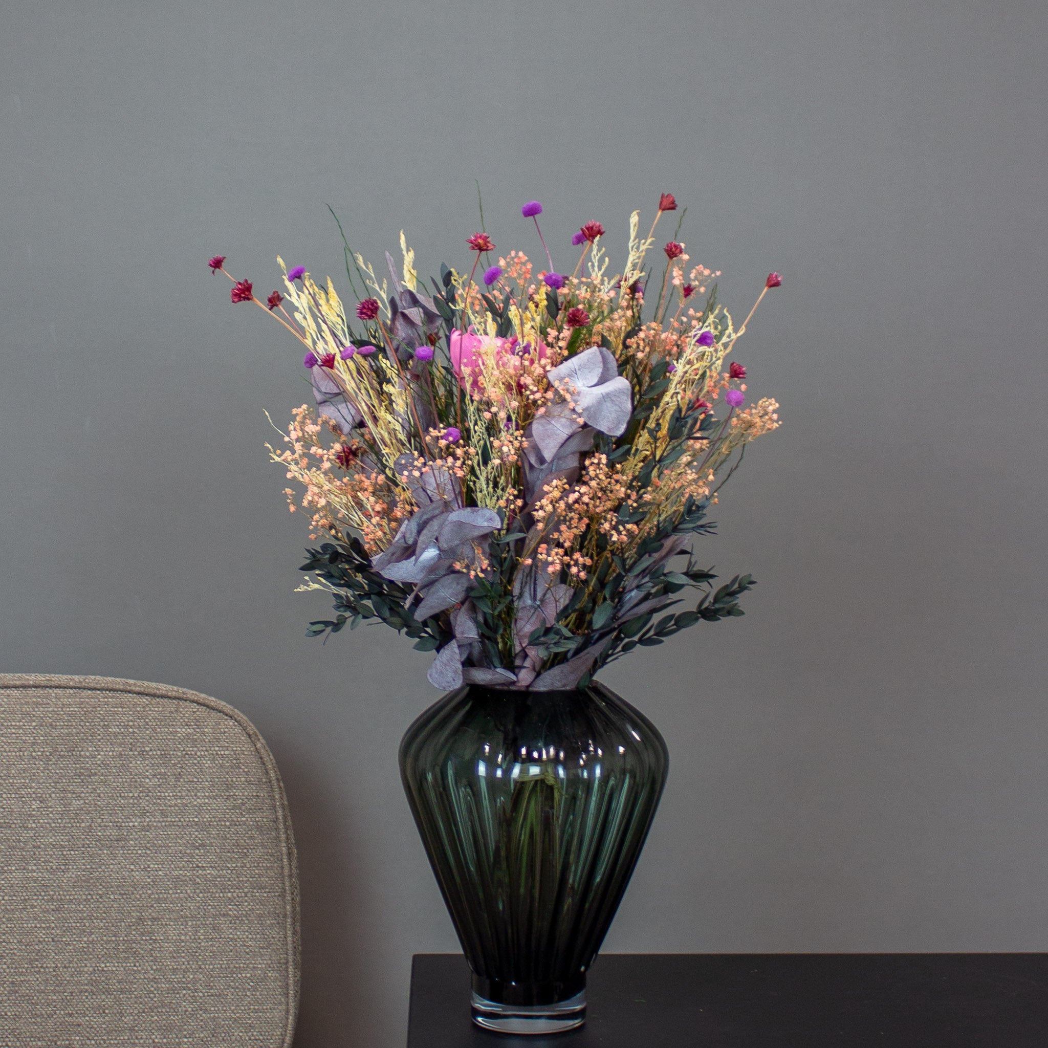 En buket i gyldne, lilla og røde nuancer står i en mørkegrøn rillet glasvase.