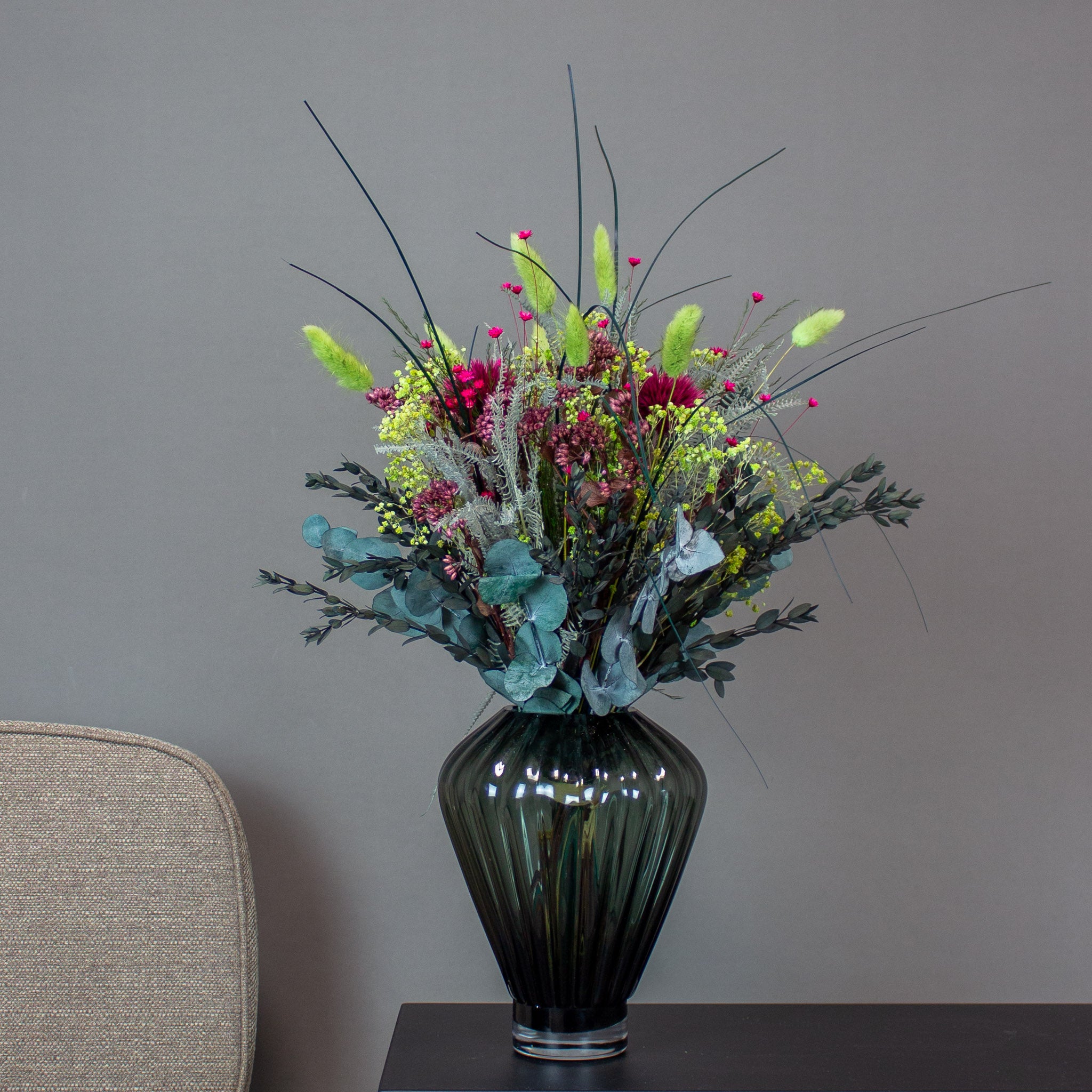 En livlig, langtidsholdbar buket i limegrønne, pink og mørkerøde nuancer står i en mørk glasvase mod en grå baggrund.