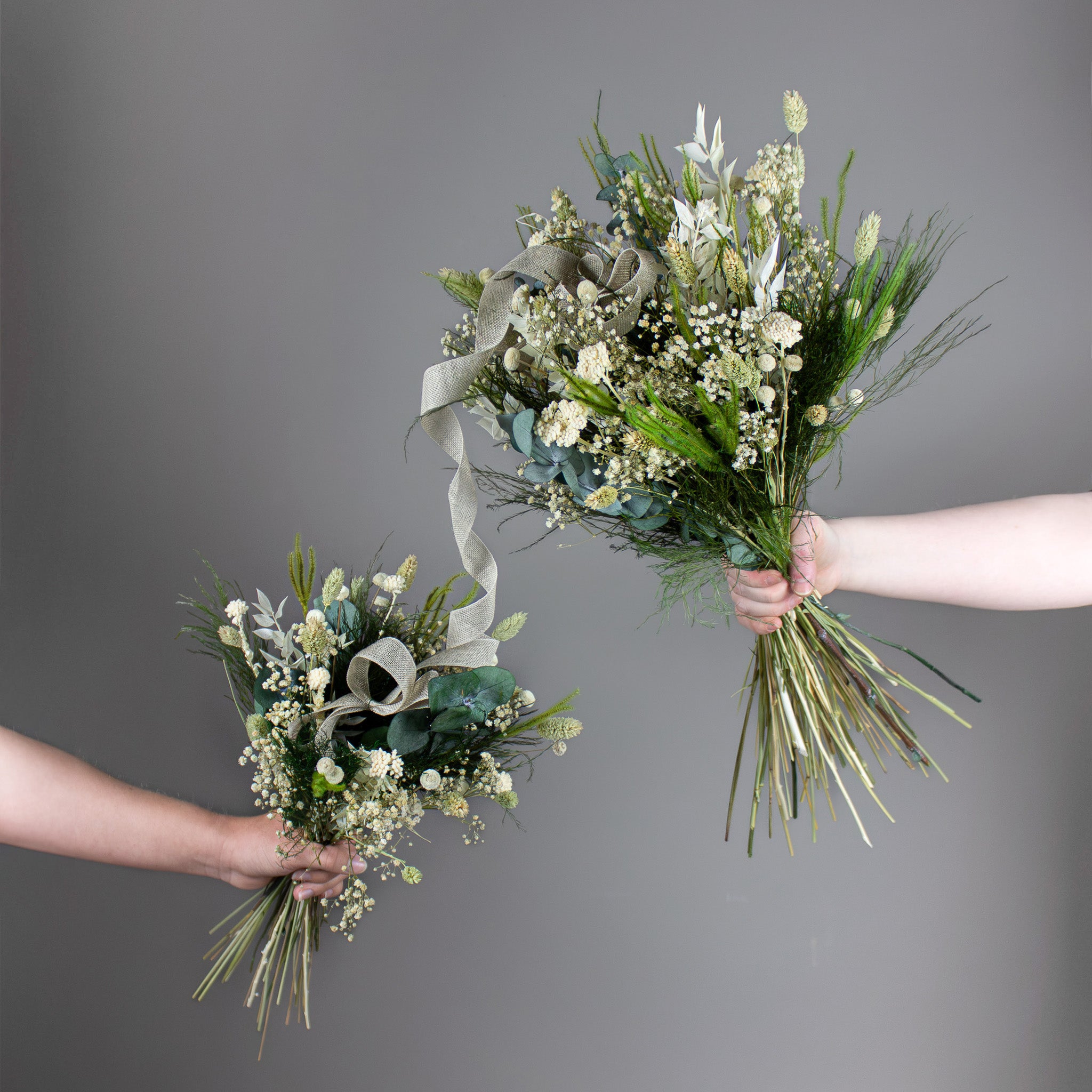 To hænder holder hver sin buket i grønne og hvide nuancer: En mindre børnebuket er forbundet med et bånd til en større buket i samme design.