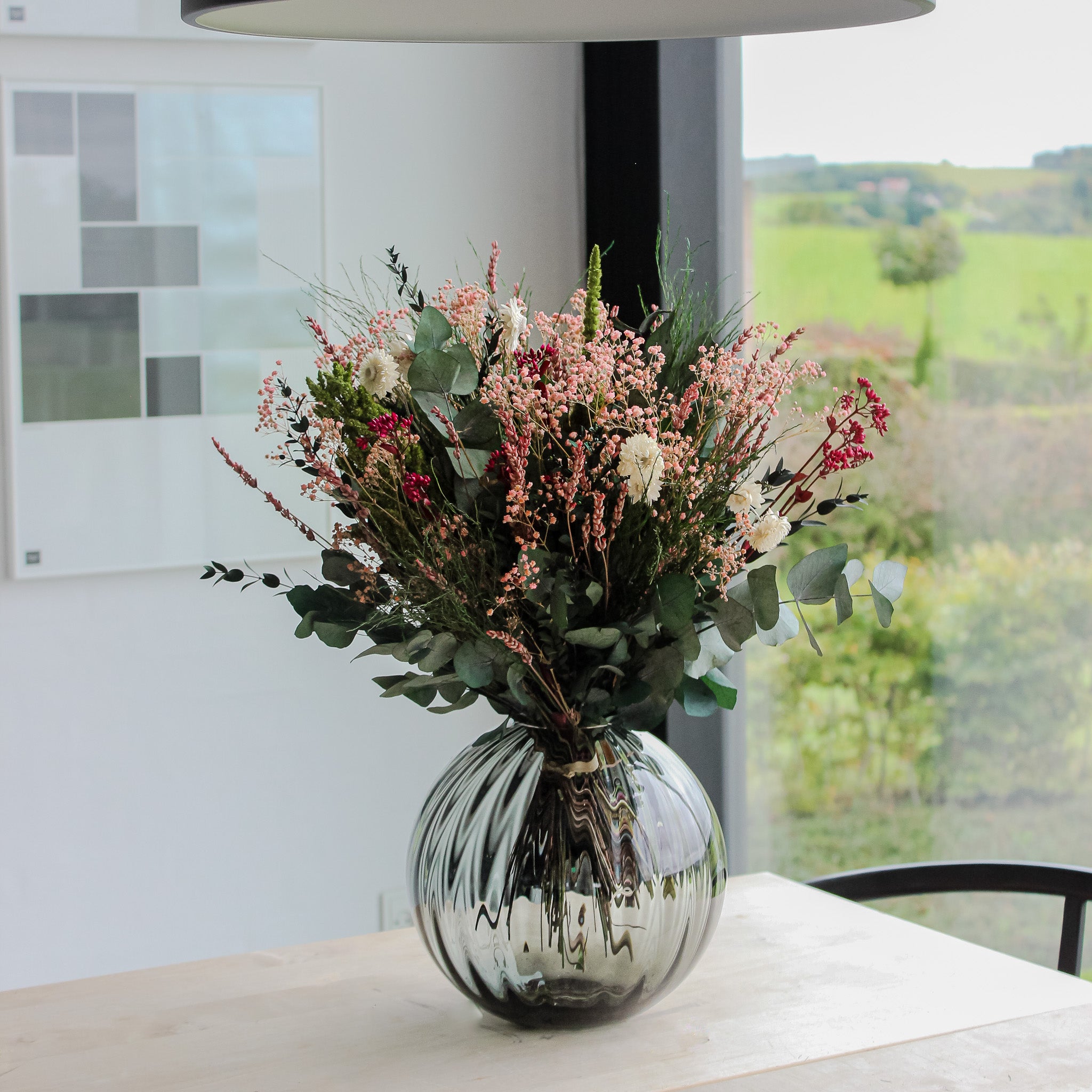 En stor, langtidsholdbar buket i rosa, creme og grønne nuancer står i en rund glasvase på et lyst træbord foran et panoramavindue.