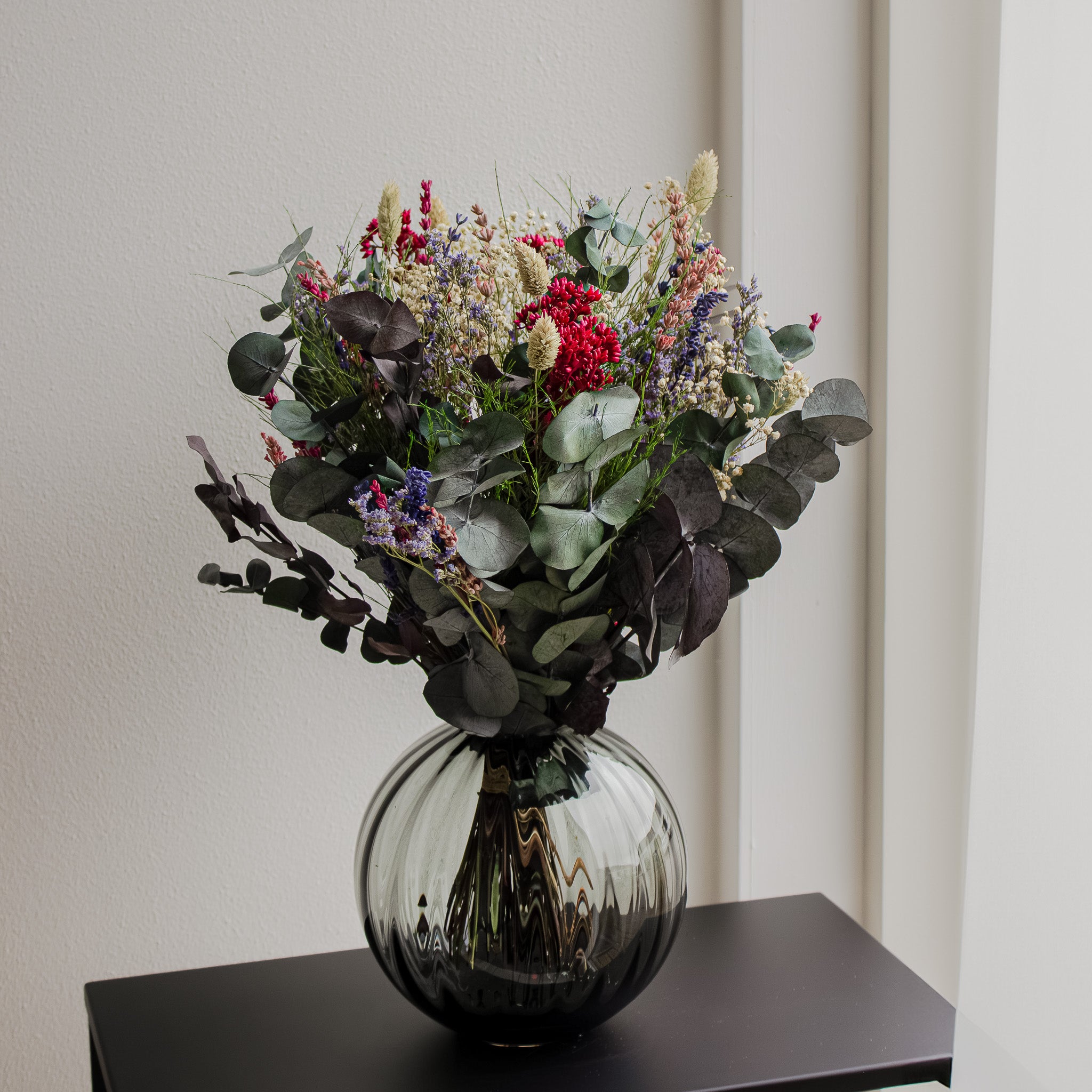 På et lille sidebord står en naturlig blomsterbuket med violette, blå og røde accenter i en buet vase.