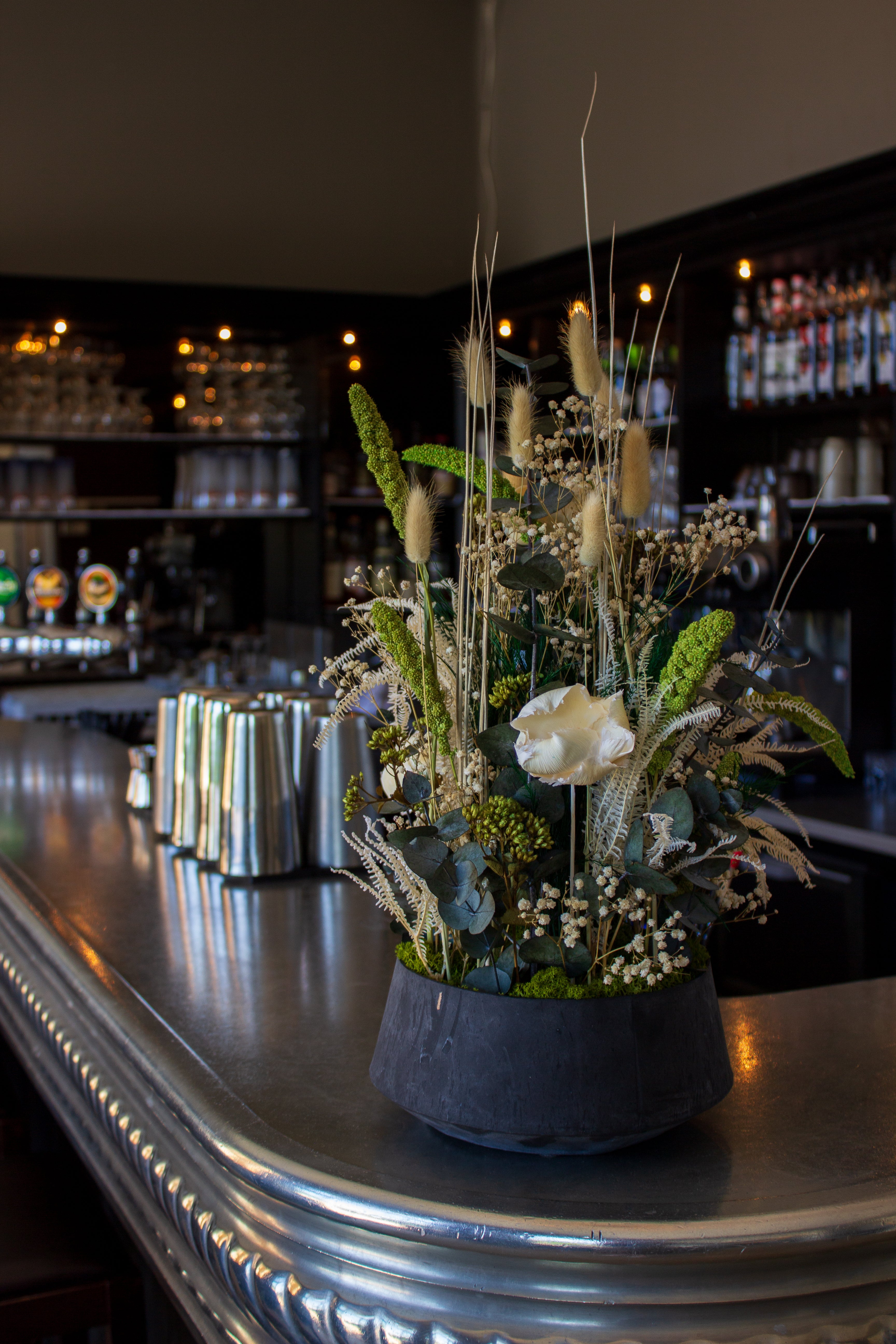 På en bardisk står en imponerende blomsterdekoration i naturlige hvid-grønne nuancer i en skål.