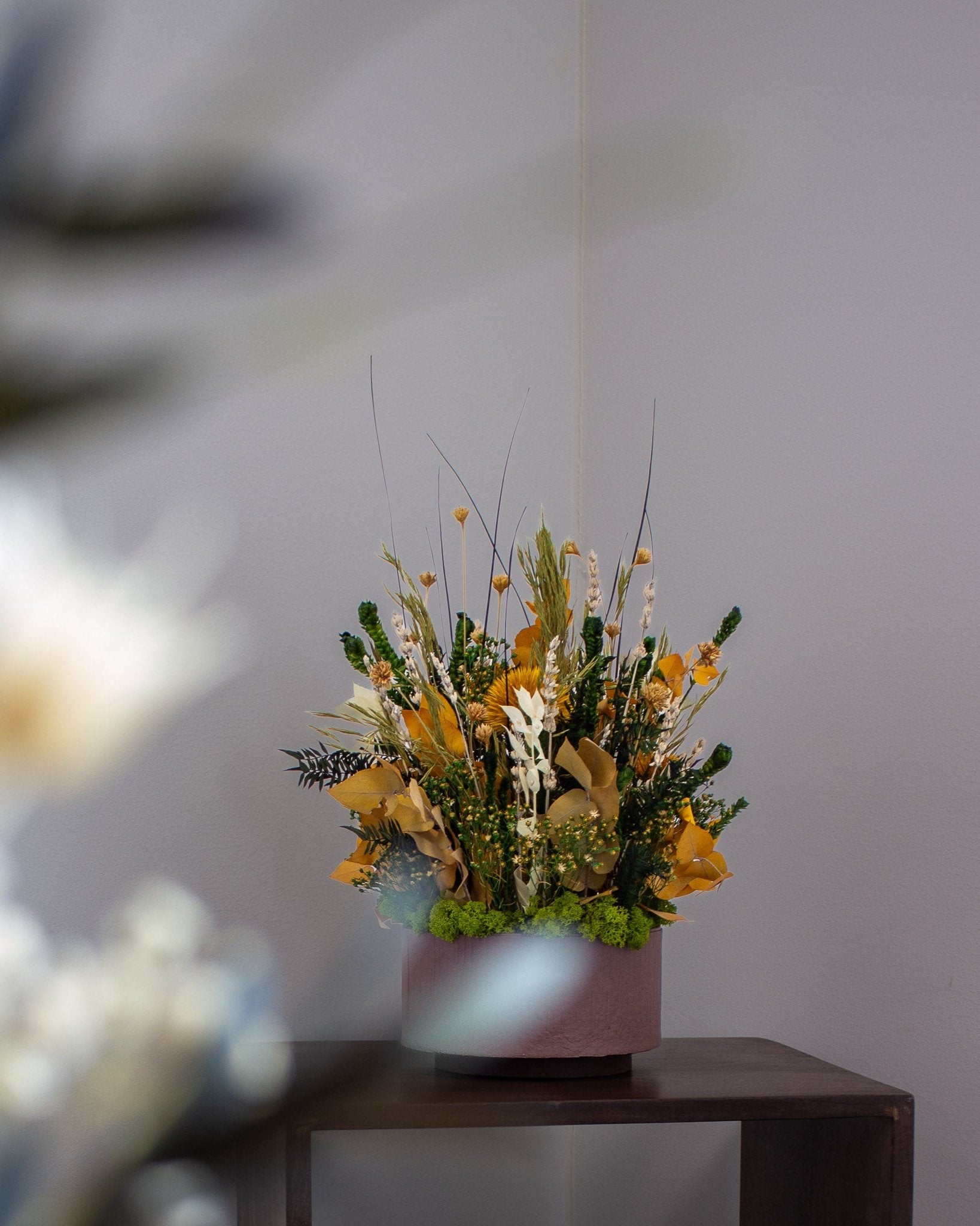 En langtidsholdbar blomsterbuket i efterårsfarver med gule, grønne og naturfarvede toner står i en lyserød beholder på et mørkt træbord foran en grå væg.